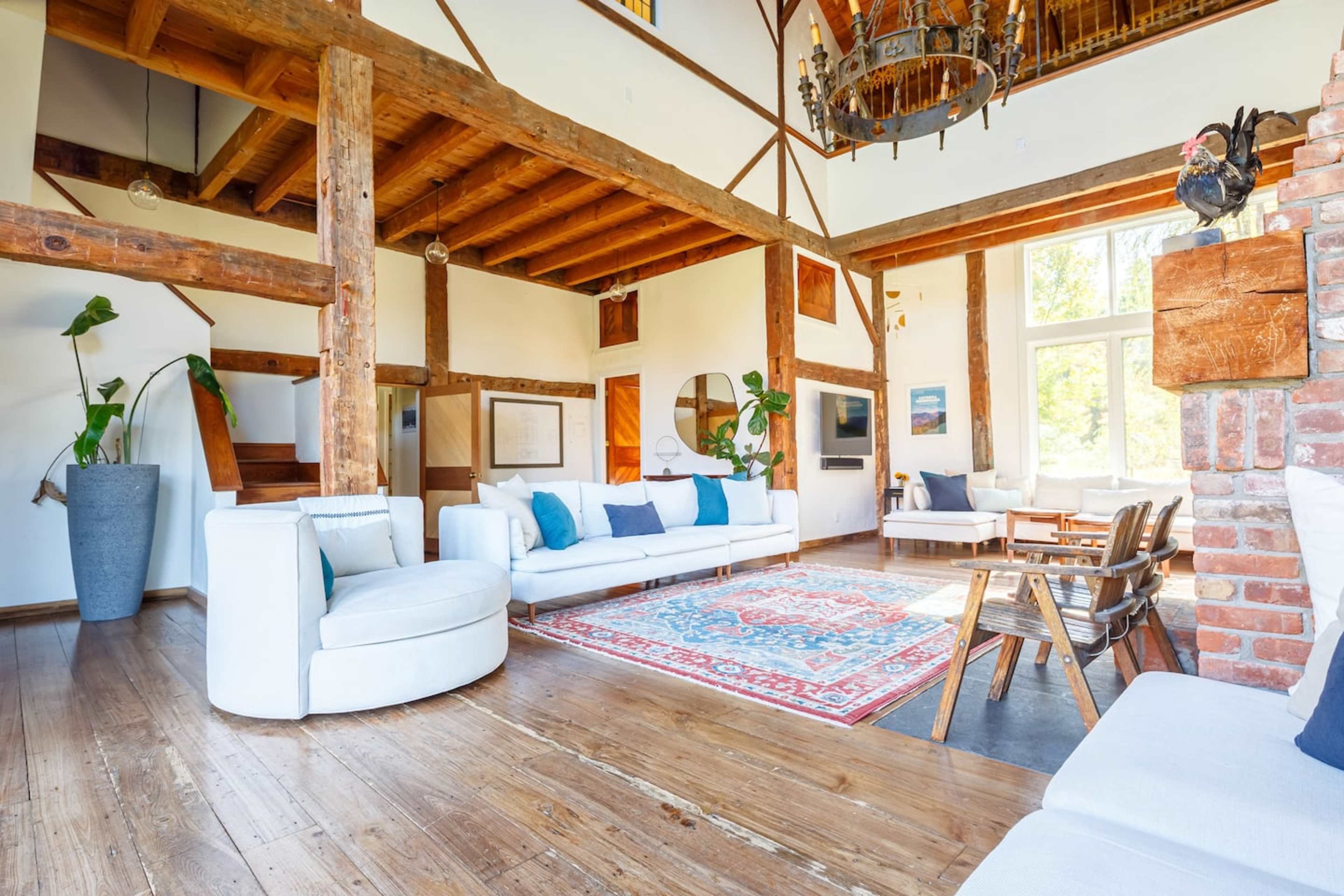 A spacious, well-lit living room featuring wooden beams, a large area rug, a mix of modern and rustic furniture, and large windows.