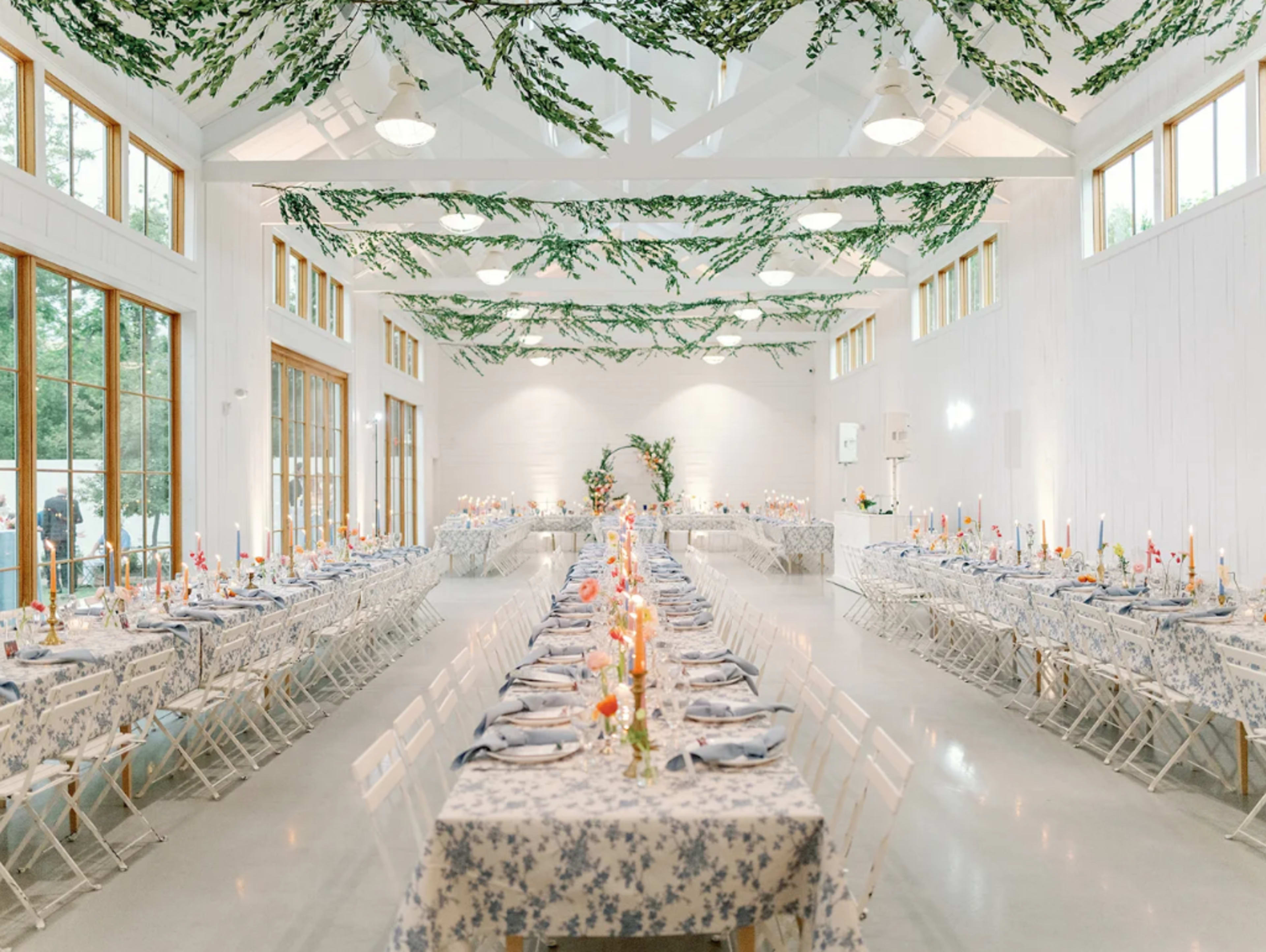 The image shows a well-decorated banquet hall featuring long tables adorned with floral tablecloths, candles, and centerpieces, while greenery is draped across the ceiling.