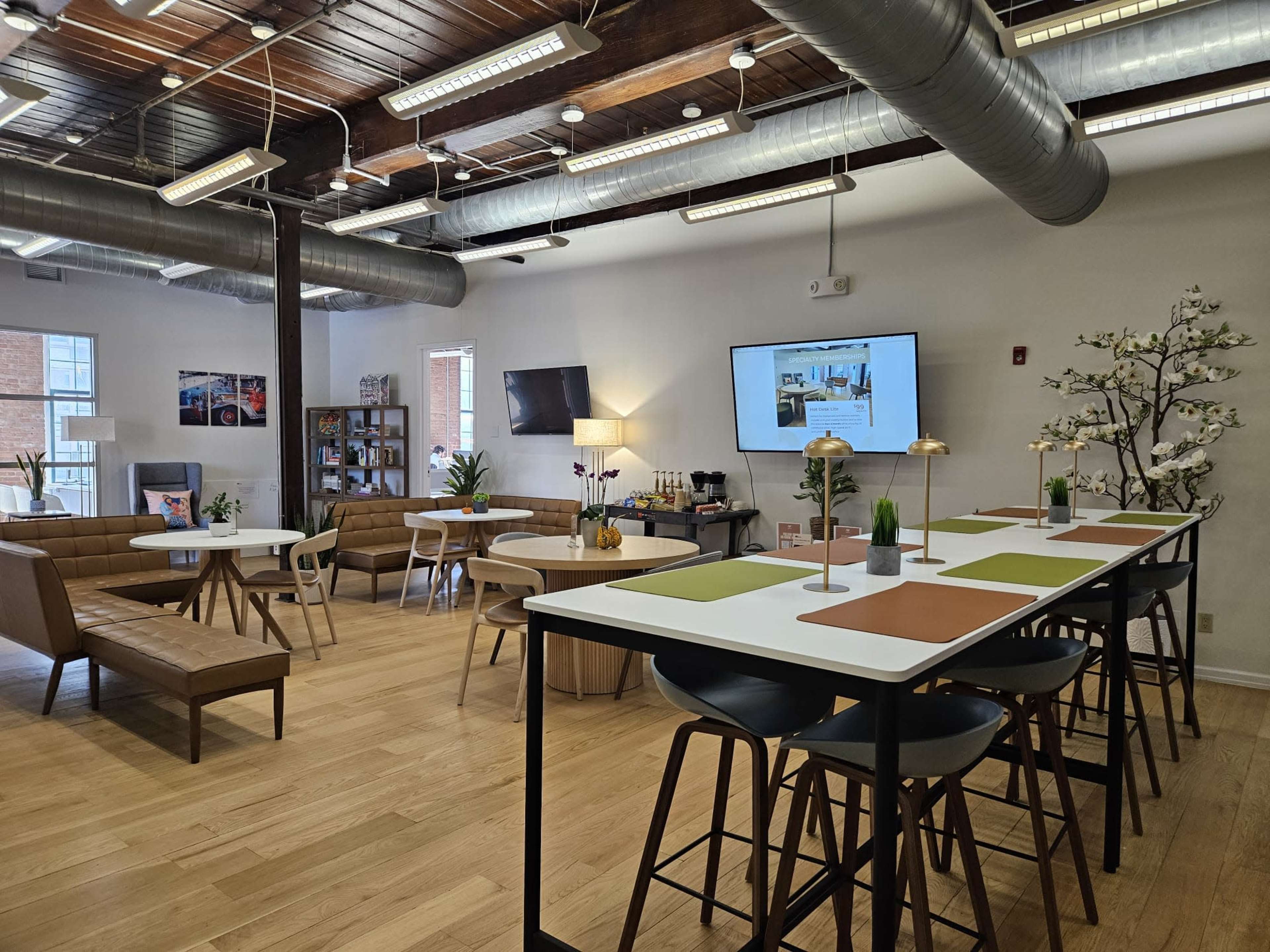 The image shows a spacious, well-lit communal area featuring seating arrangements, a large screen, and dining tables with greenery and decorative elements.