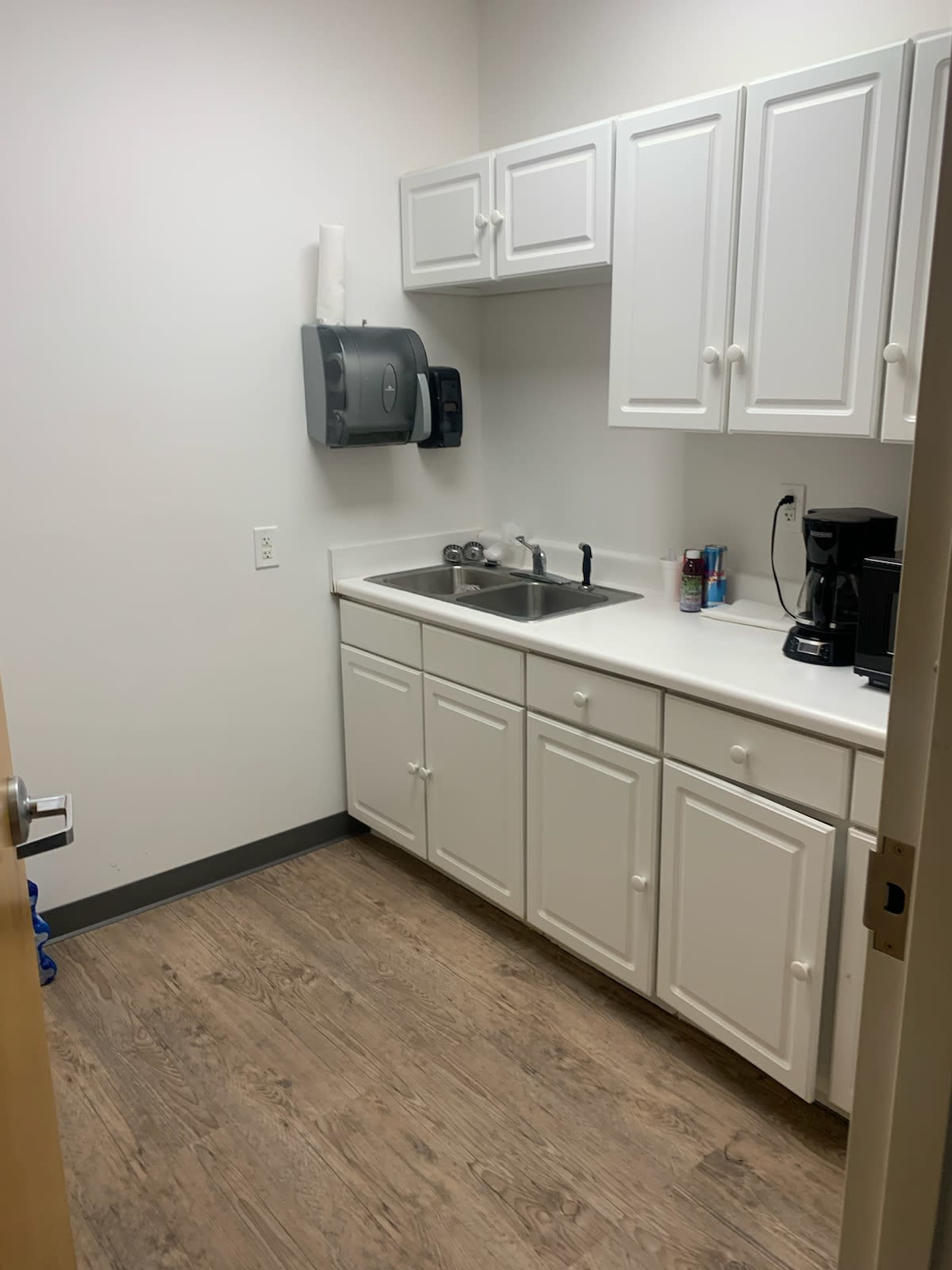 A small kitchenette with white cabinets, a sink, and a coffee maker against a plain wall.