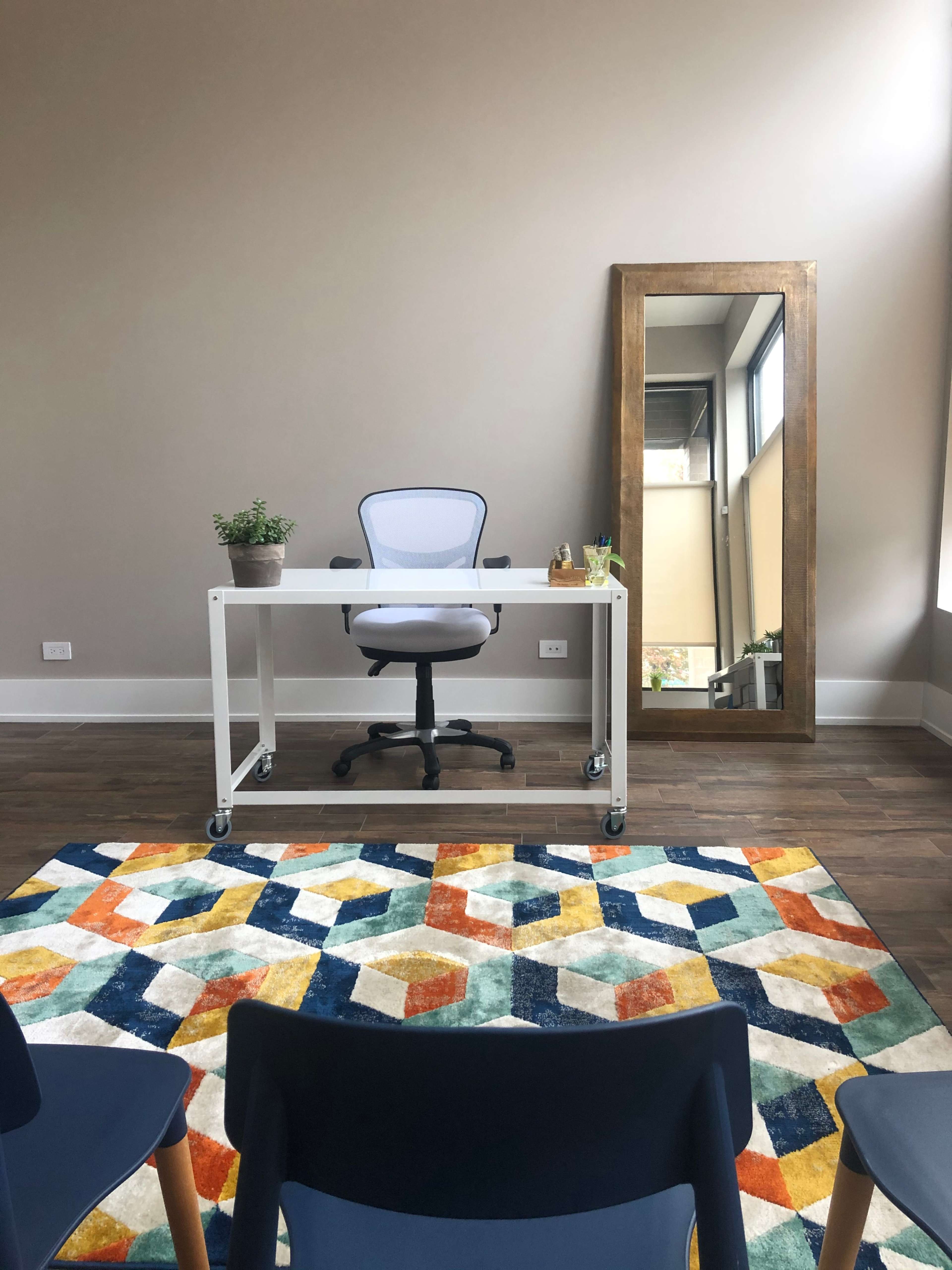 A minimalist office space featuring a white desk and chair in front of a large mirror, with a colorful geometric rug on the wooden floor.