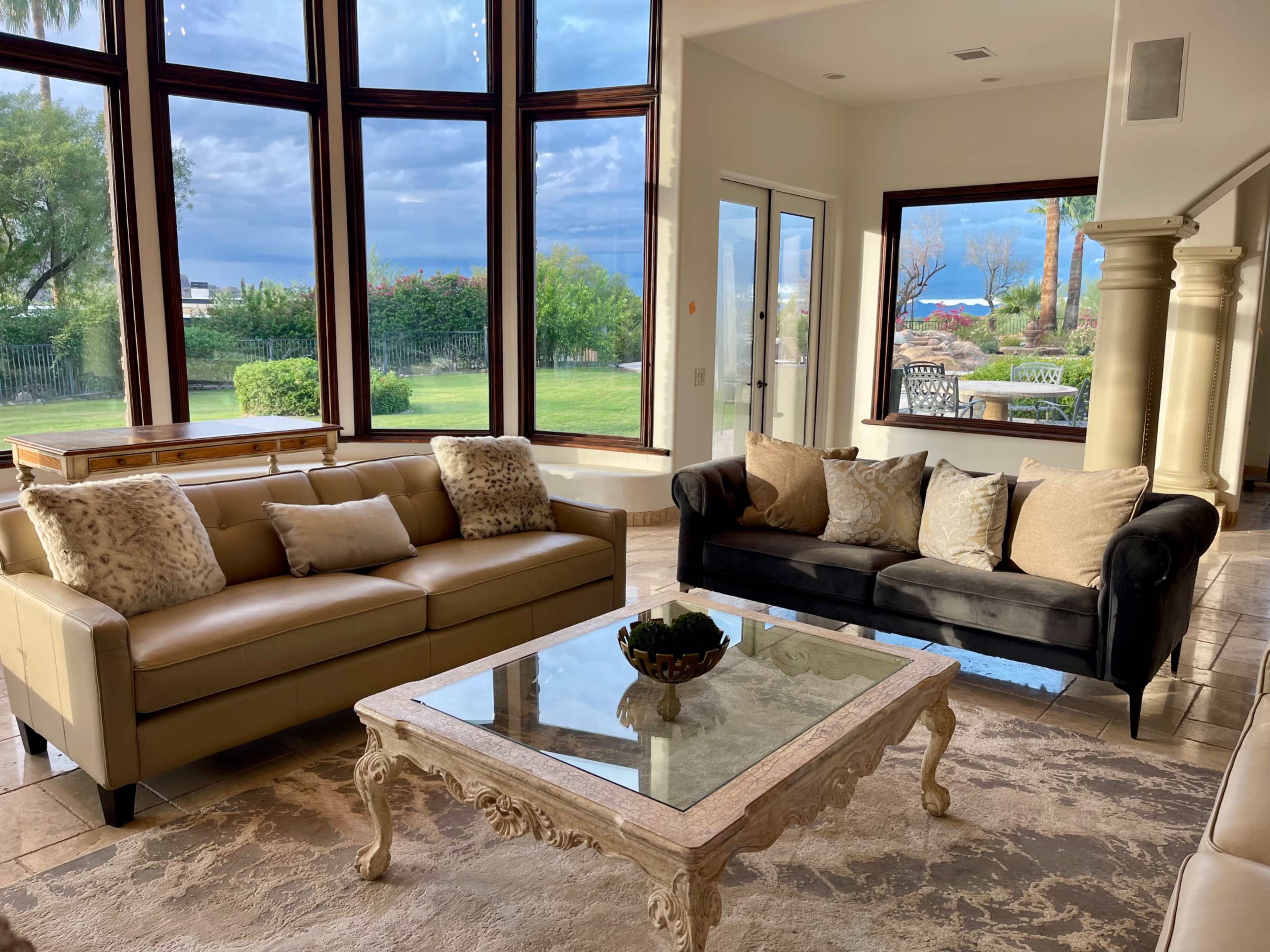 The image shows a spacious living room with large windows, featuring two sofas, a coffee table, and a view of greenery outside.