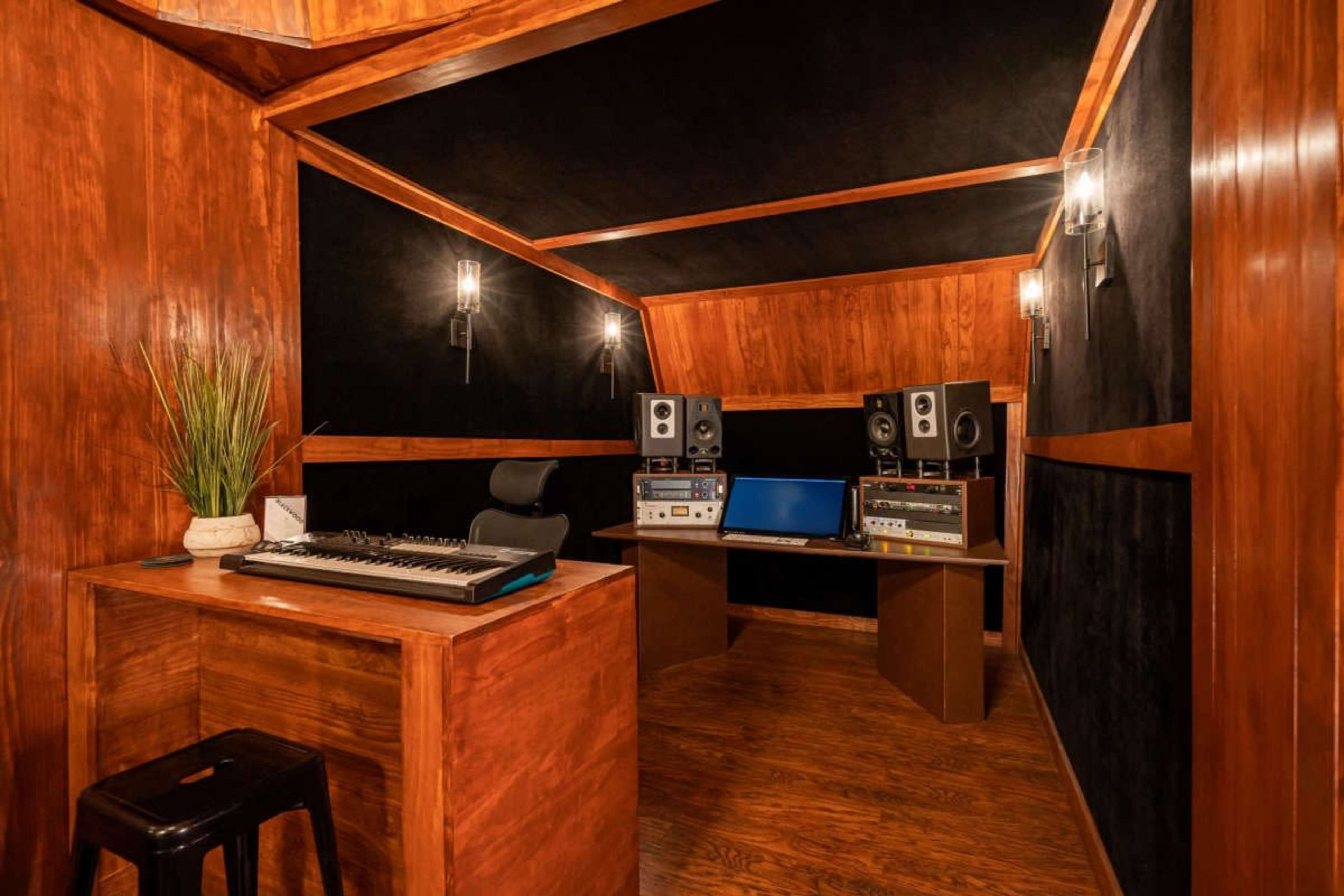 The image shows a small music recording studio featuring wooden walls, a desk with a computer and audio equipment, a keyboard on the side, and track lighting.