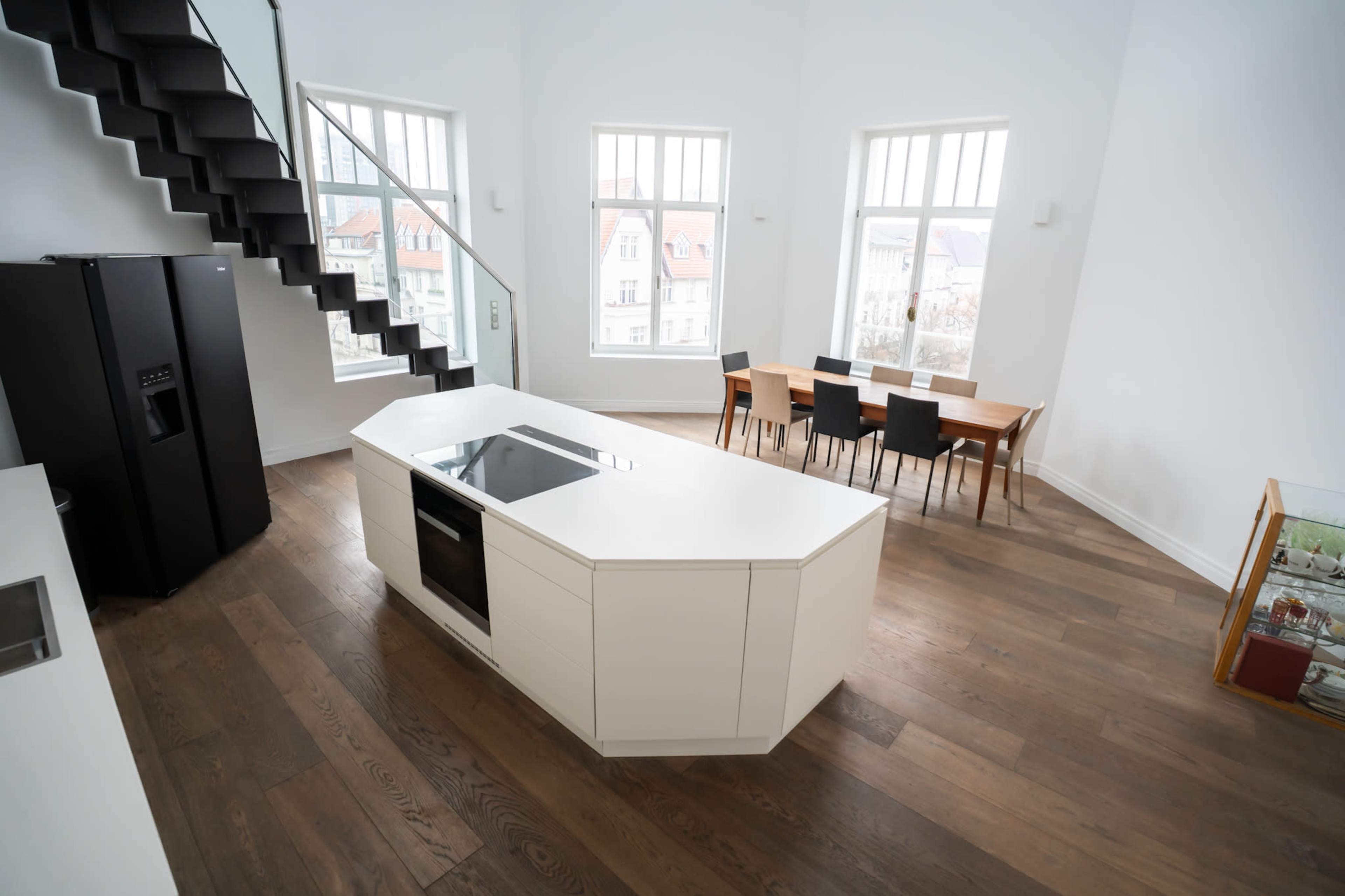 The image shows a modern kitchen with a central island, black refrigerator, and a dining table surrounded by chairs, all set in a bright, spacious room with large windows.