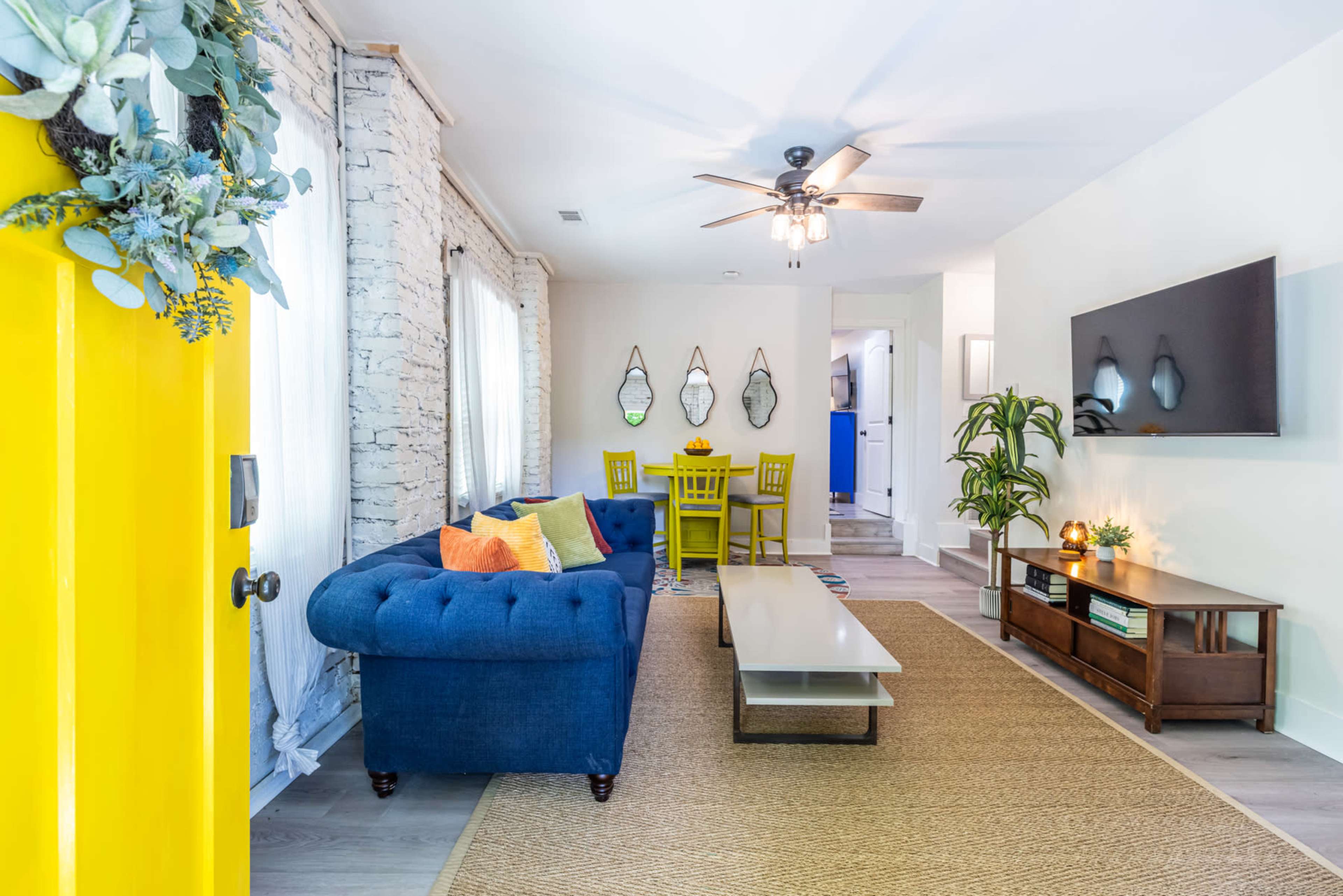 The image shows a bright, modern living room with a blue sofa, a wooden coffee table, and a dining area with yellow chairs in the background.