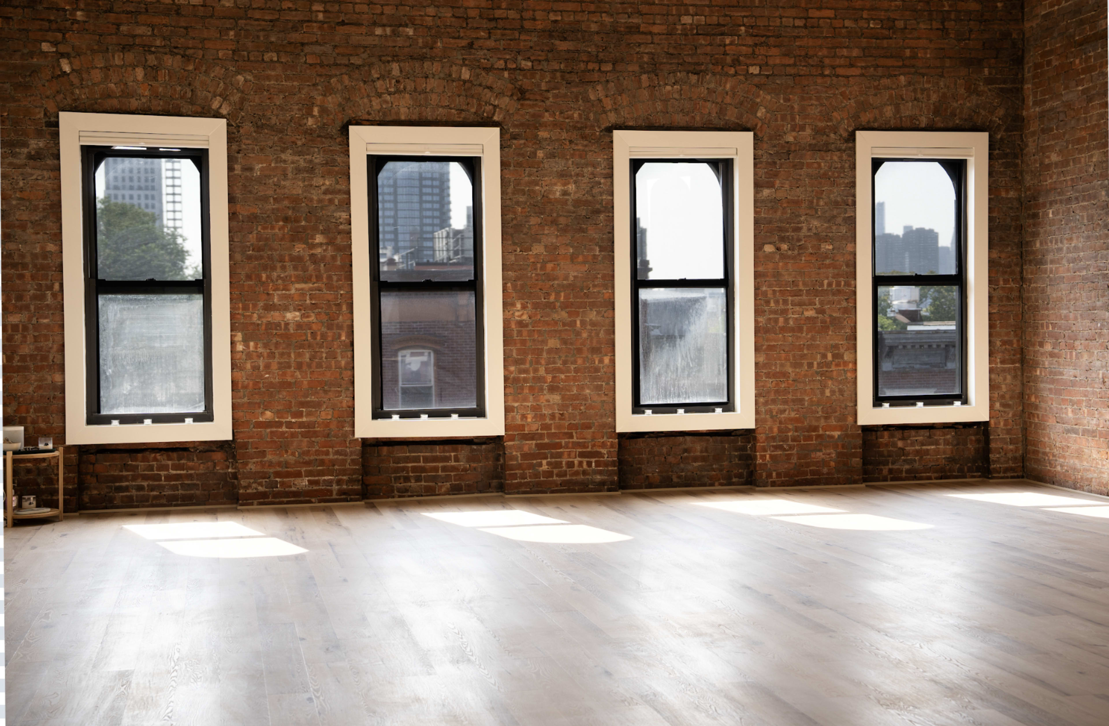 The image shows an empty room with four tall windows along a brick wall, allowing natural light to illuminate the wooden floor.