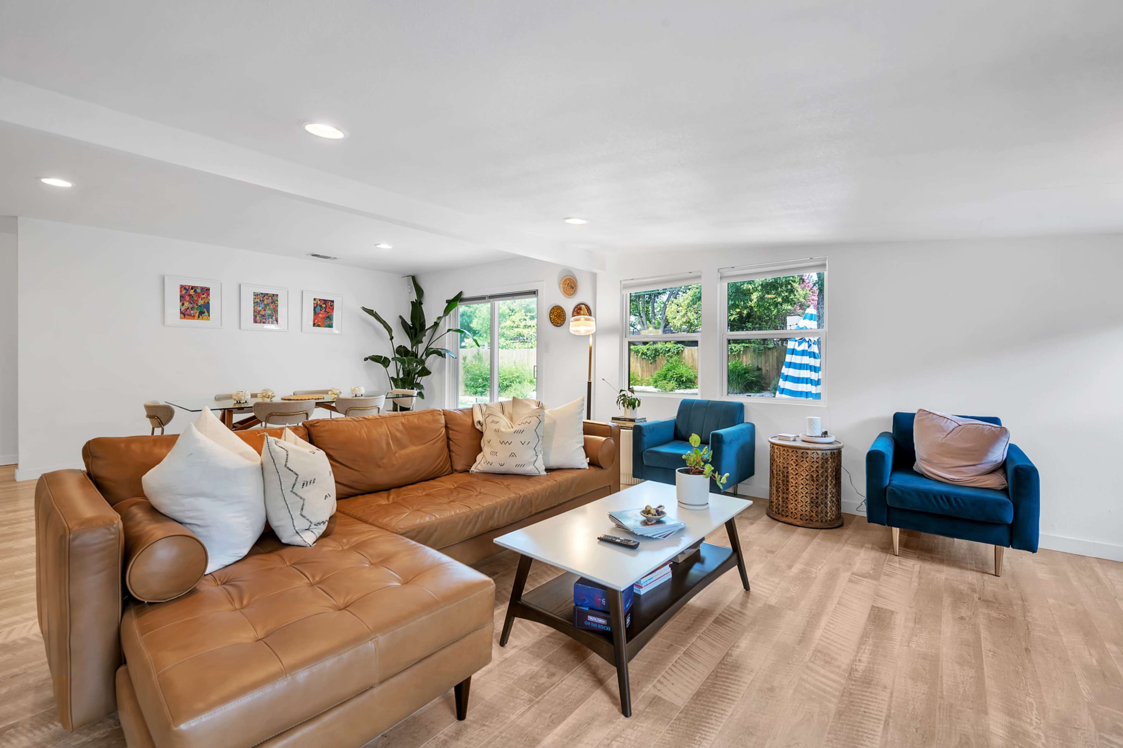 The image shows a bright and spacious living room featuring a brown leather sectional sofa, a blue armchair, a coffee table, and a dining area with a large window overlooking greenery.