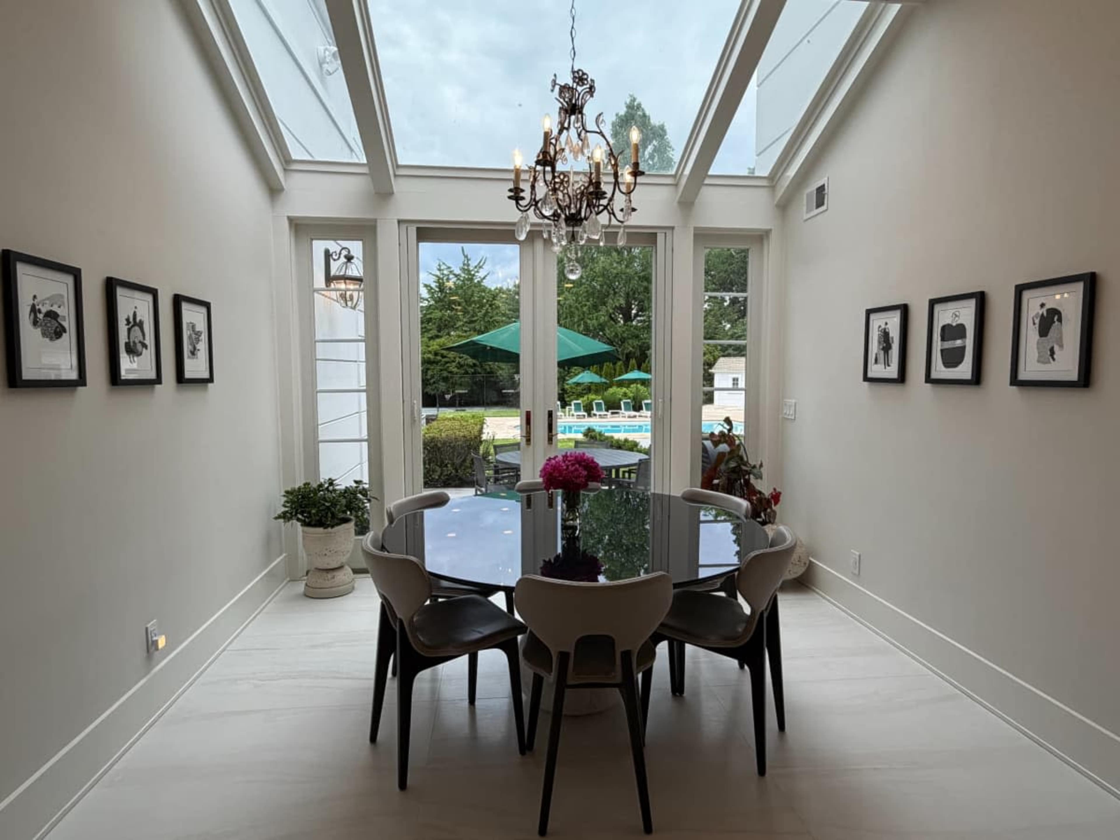A dining room features a glass table surrounded by six chairs, with large windows allowing natural light and a view of an outdoor pool area.