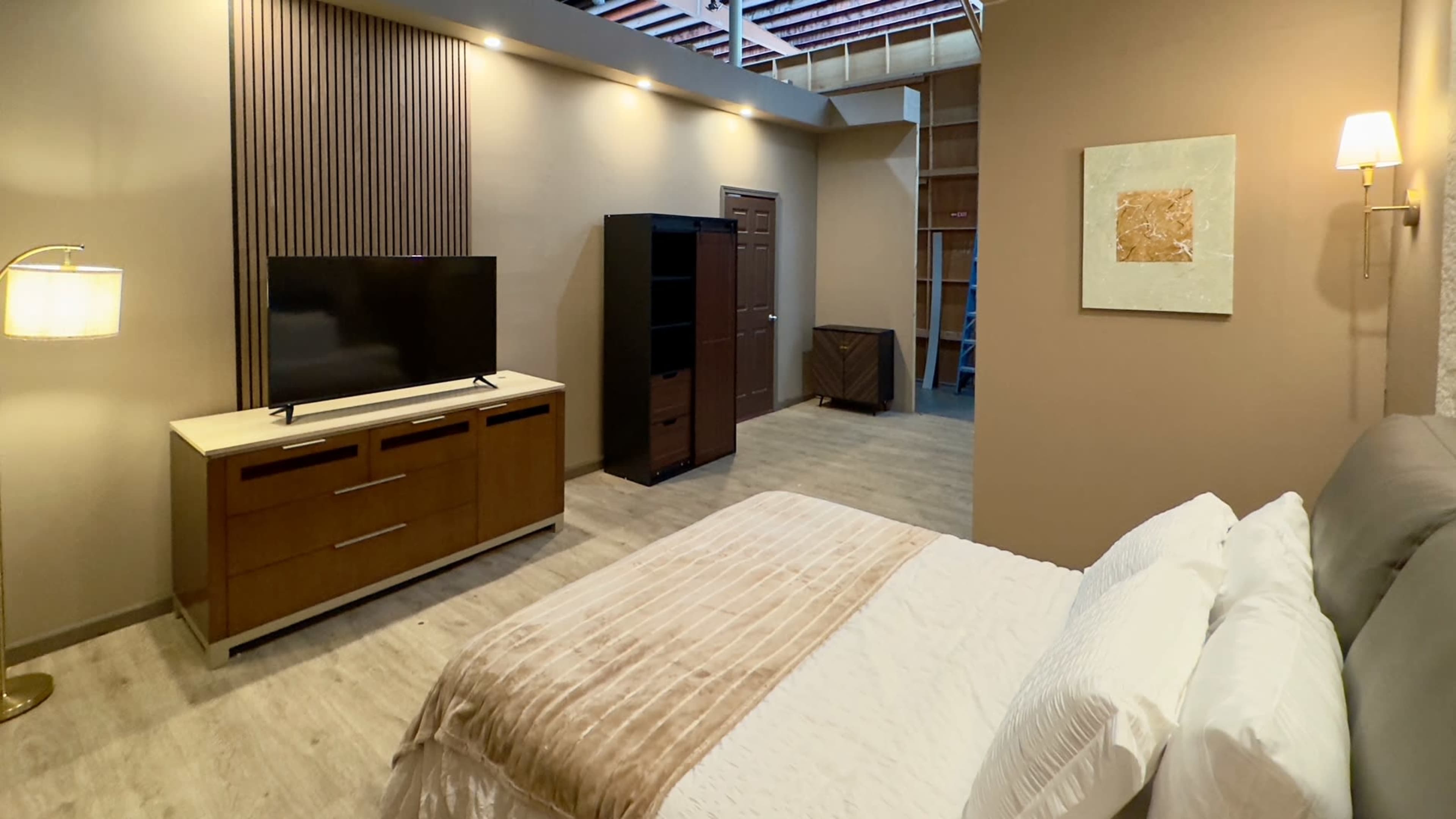 A modern bedroom with a bed, a flat-screen TV on a dresser, a bookshelf, and a door leading to another room.