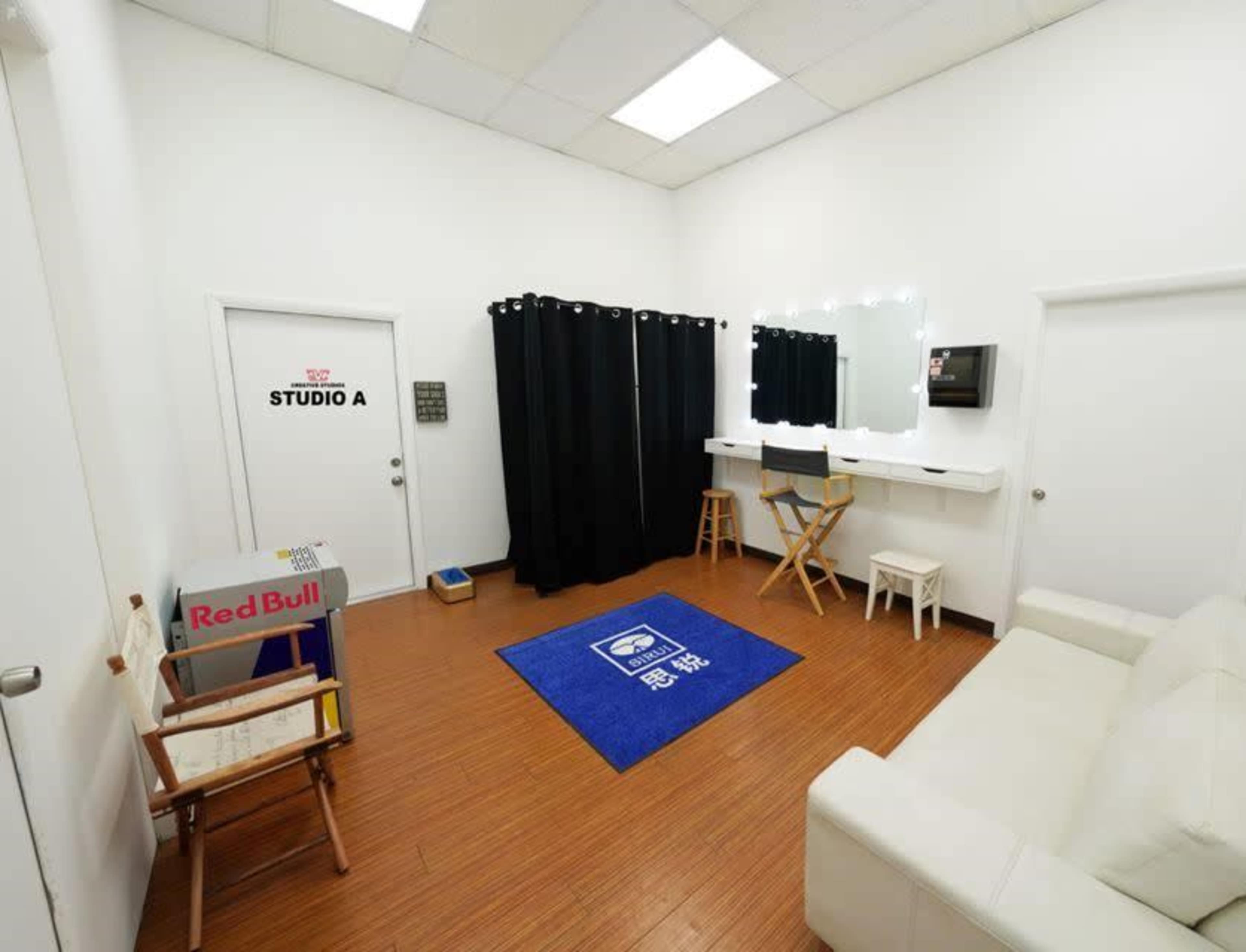 A small room with a white interior featuring a couch, a dressing area with mirrors and makeup lights, a director's chair, and a Red Bull box.