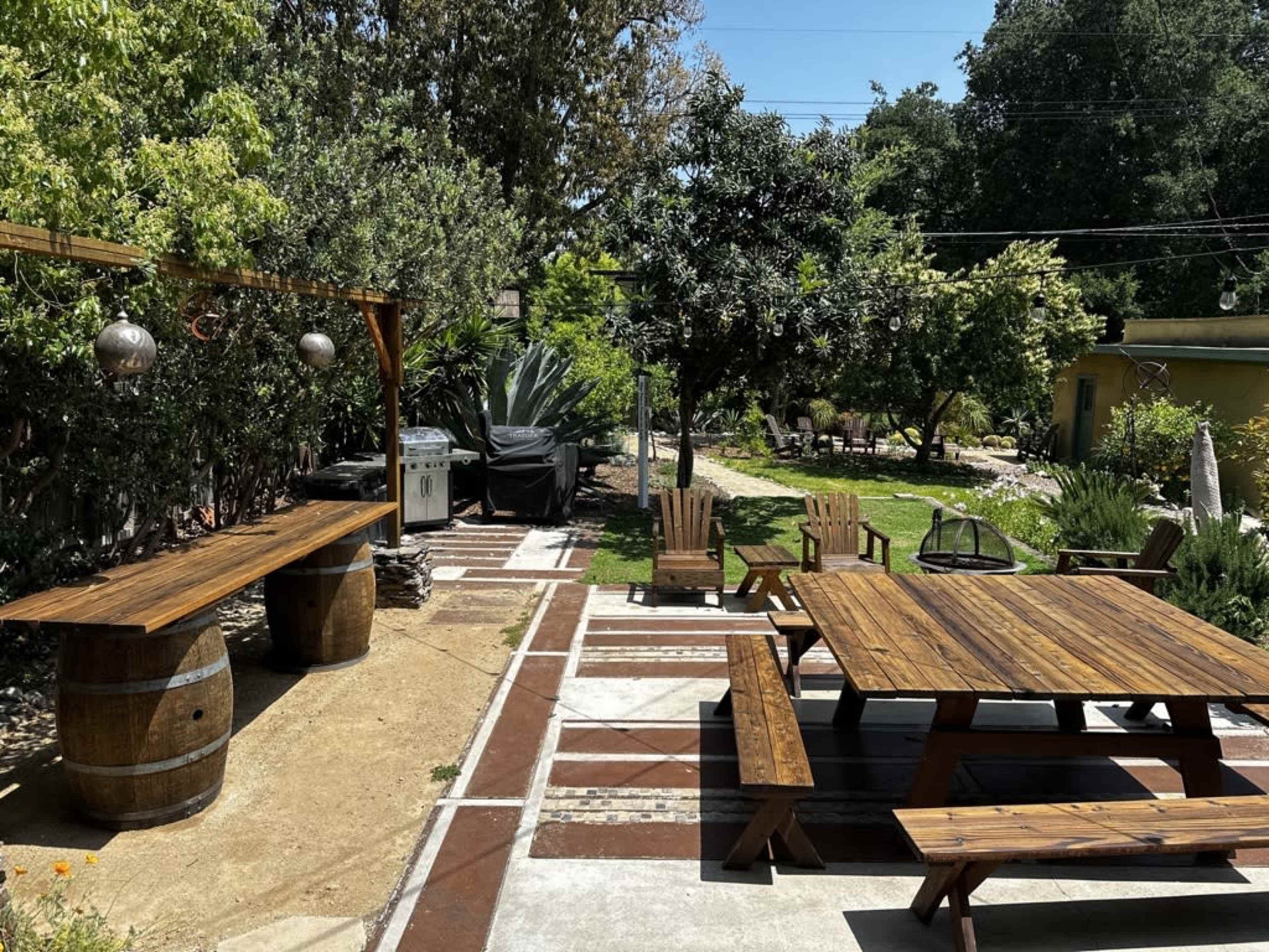 A garden area with wooden picnic tables, benches, and barrels, surrounded by greenery and a barbecue grill in the background.