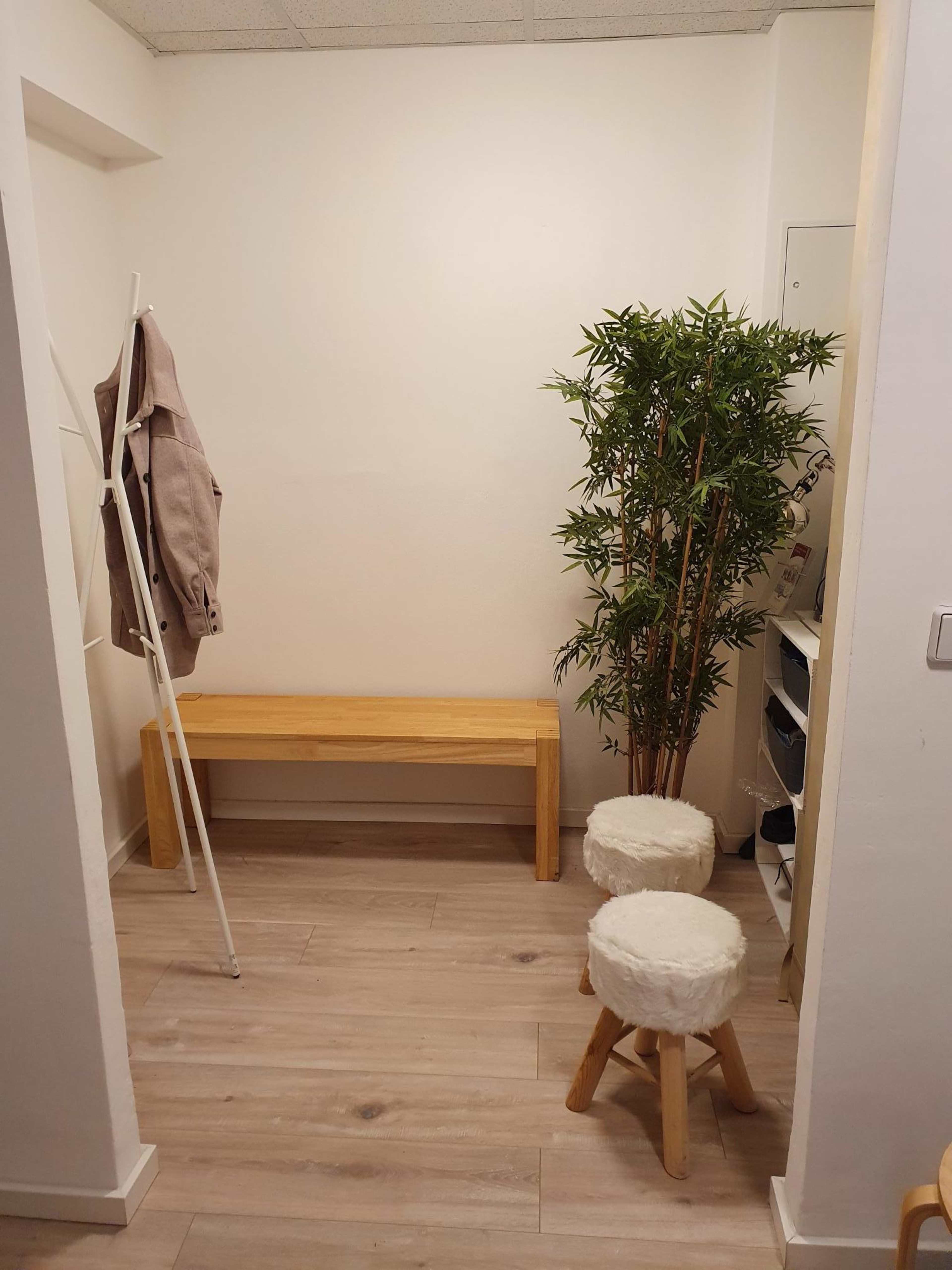 The image shows a small, minimalist room with a wooden bench, two round stools, a potted plant, and a coat rack against a white wall.