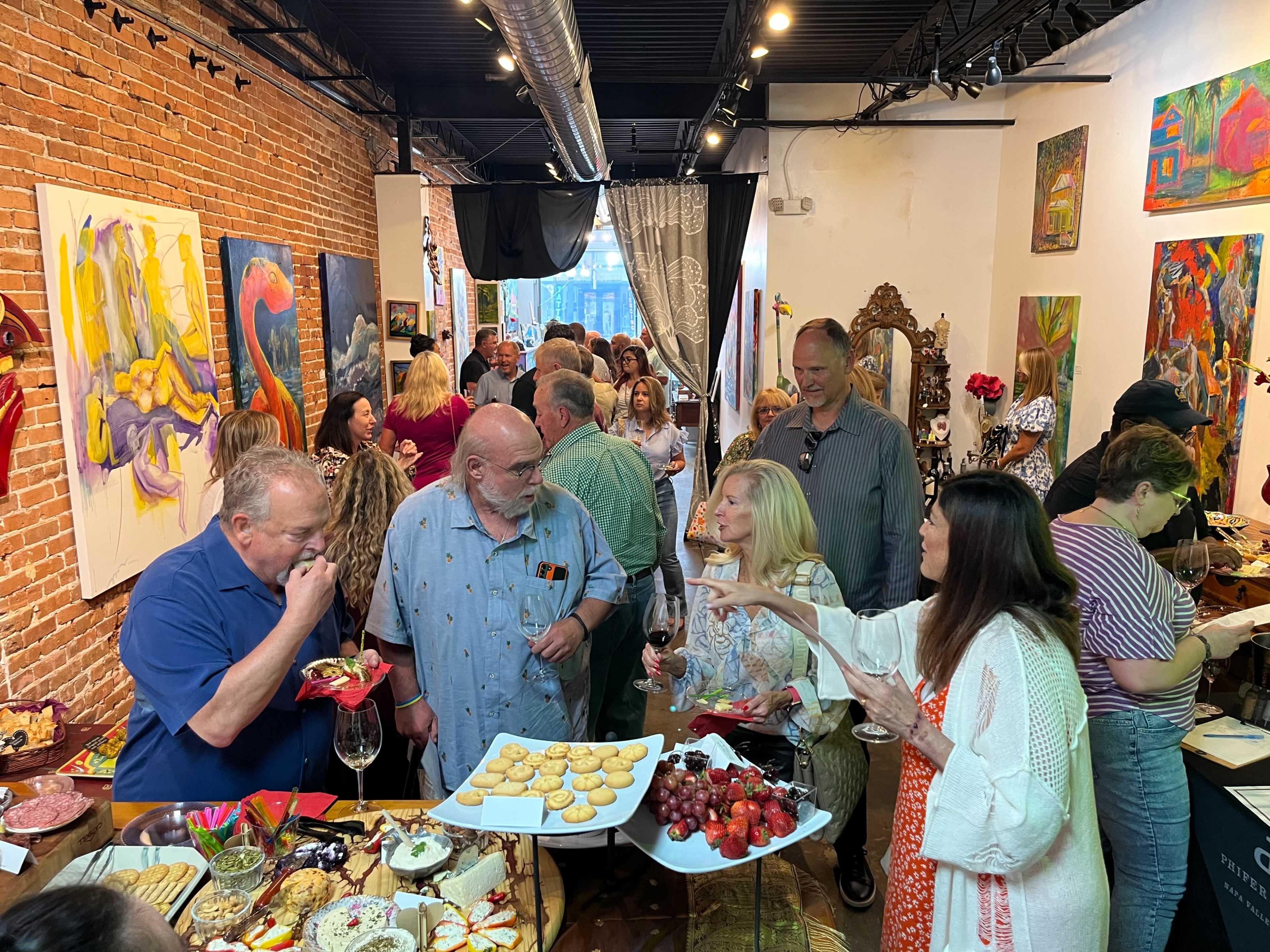 A group of people socializes while enjoying a buffet in an art gallery filled with colorful paintings.