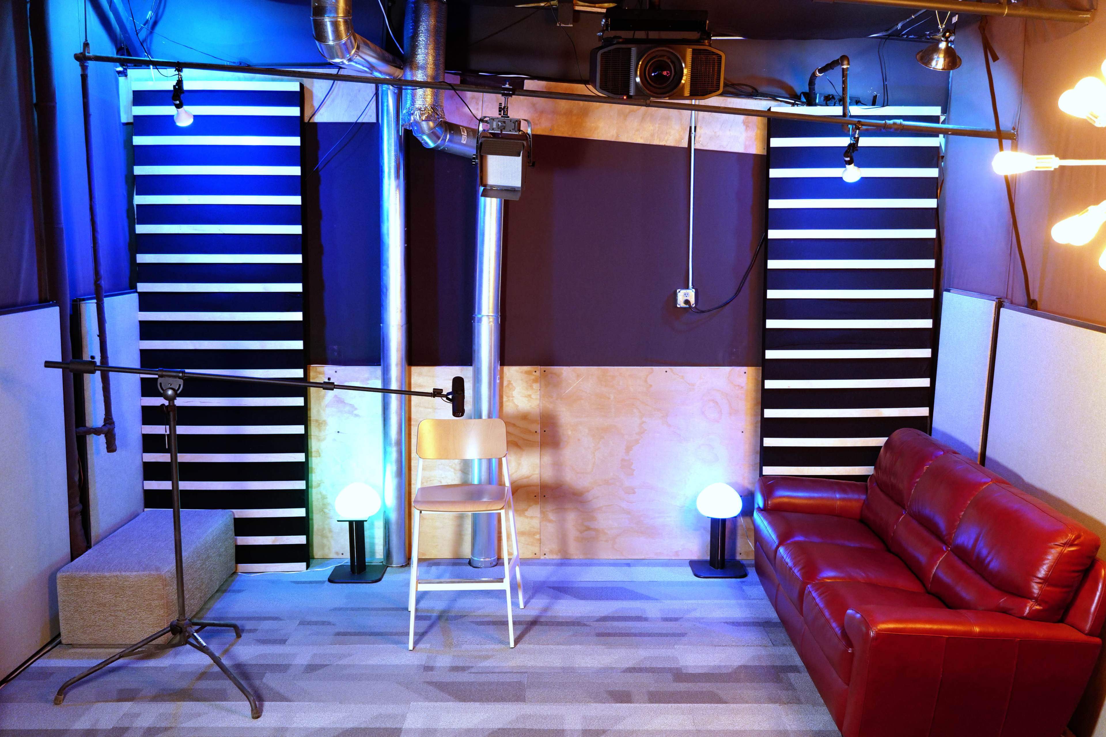 A small recording or interview studio featuring a red couch, a wooden chair, and two floor lamps, with soundproofing panels and microphone equipment positioned for use.