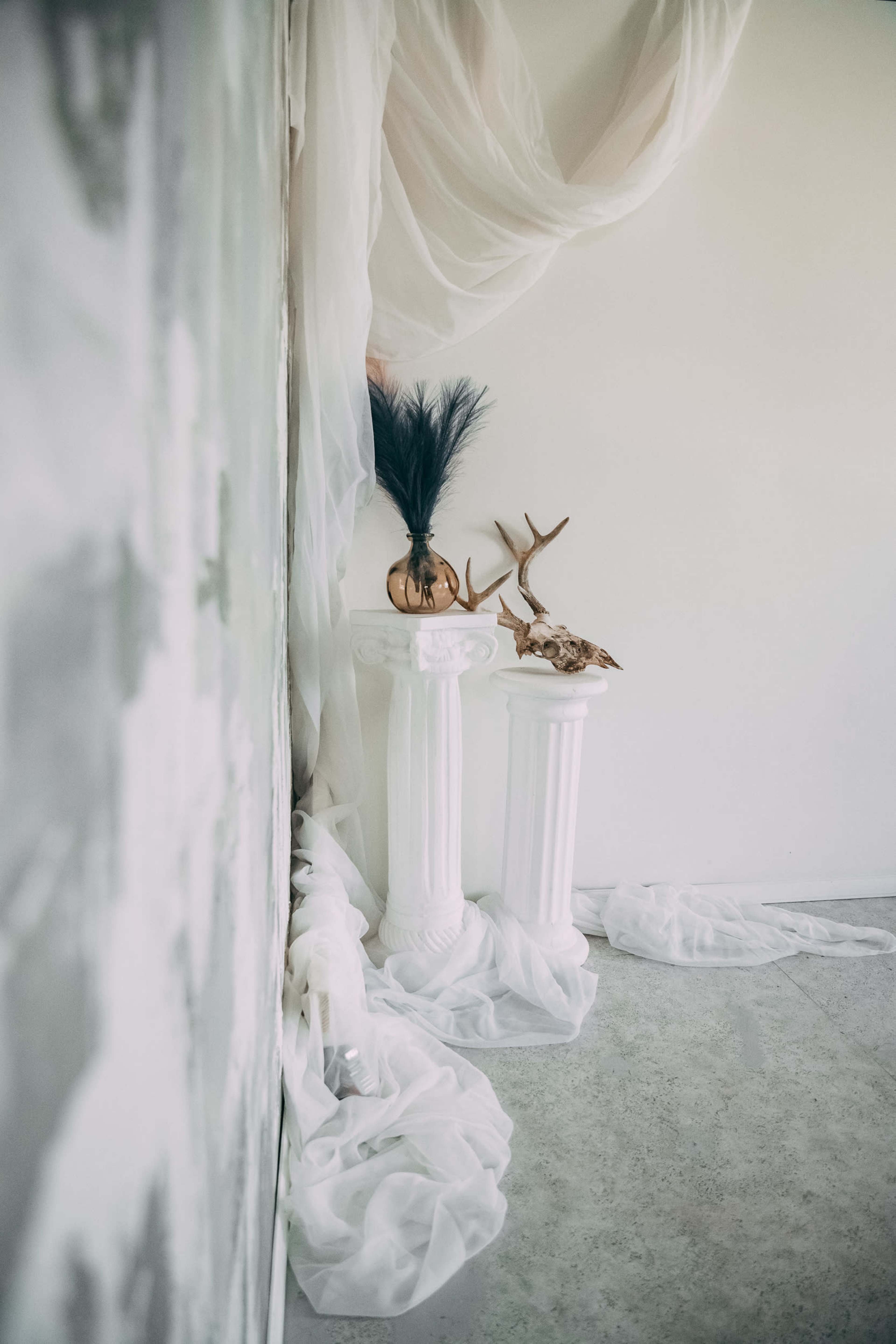 The image features two white pillars adorned with decorative items, including a vase with feathers and animal antlers, set against a backdrop of draped fabric and a pale wall.