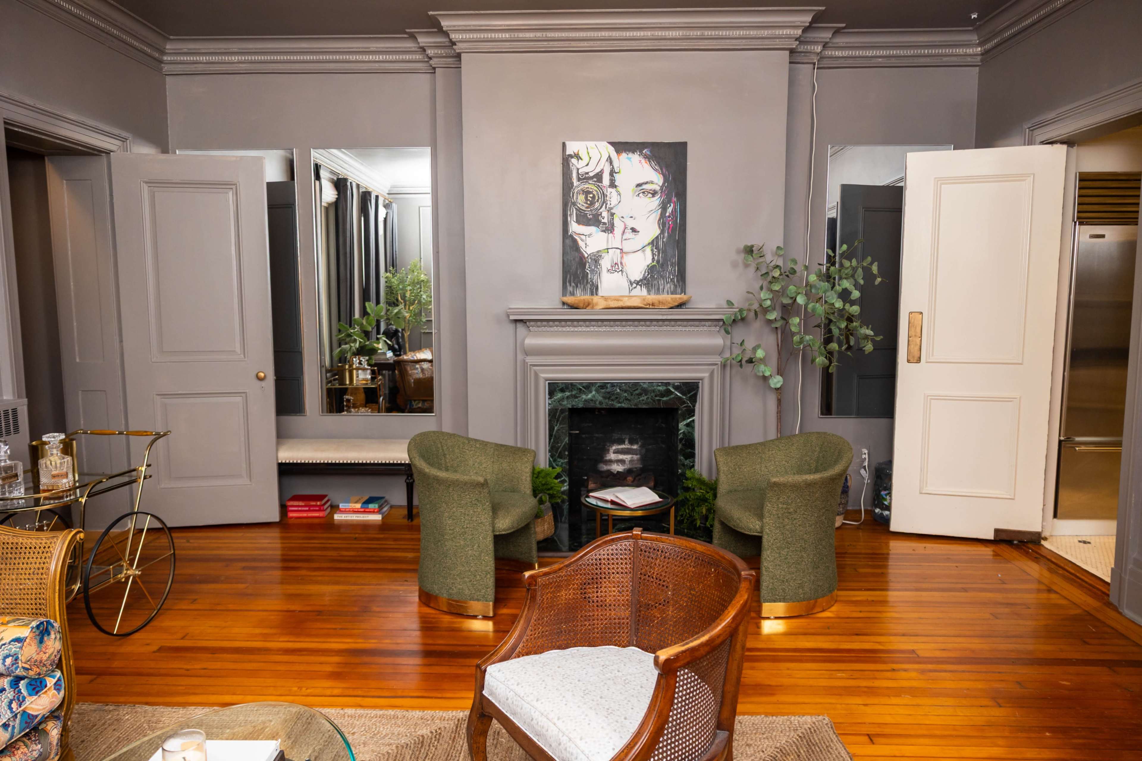 The image shows a living room featuring two green chairs, a fireplace with a painting above it, and large doors leading to another area.