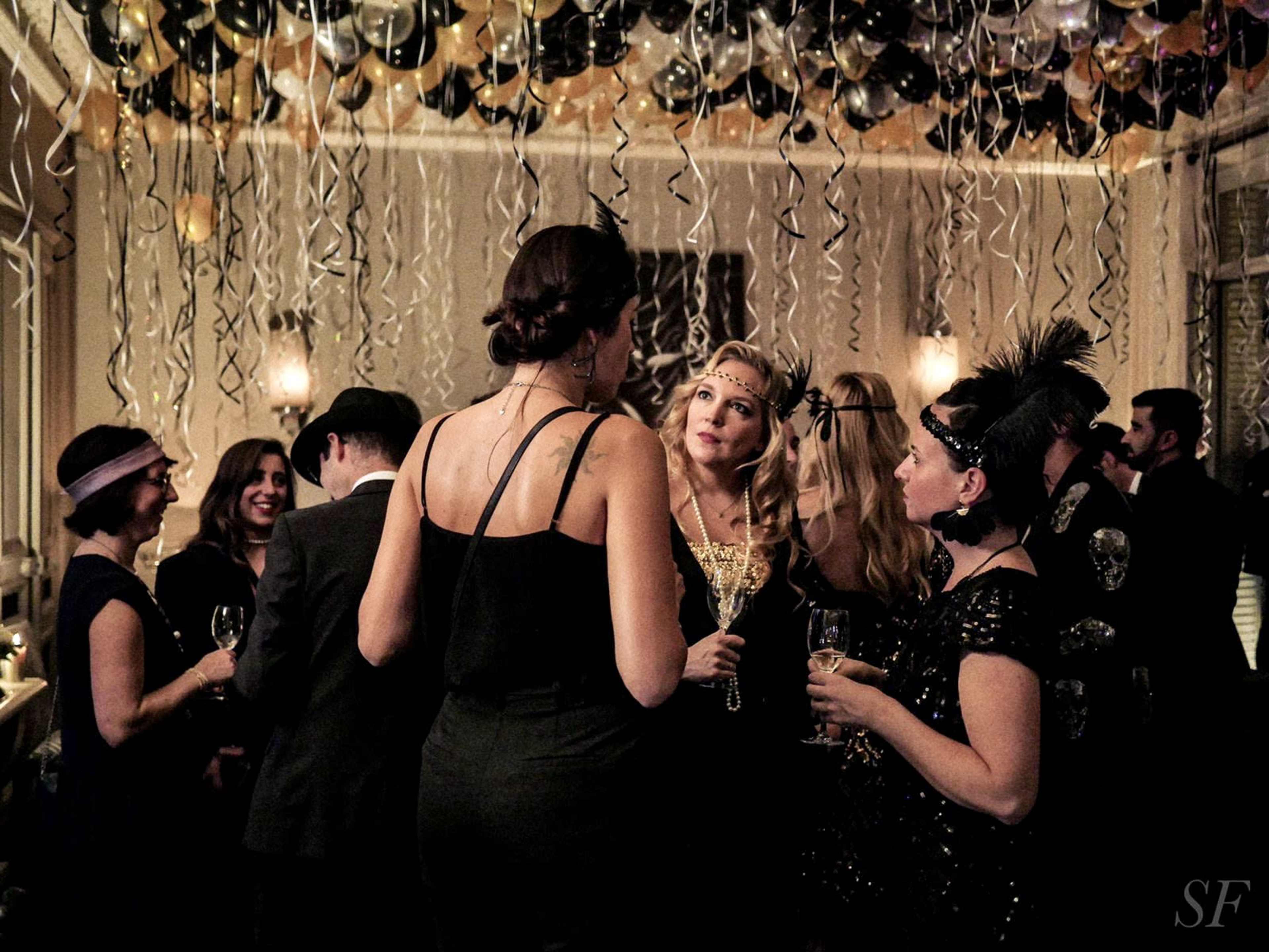 A group of elegantly dressed people mingle at a festive gathering decorated with black and gold balloons.