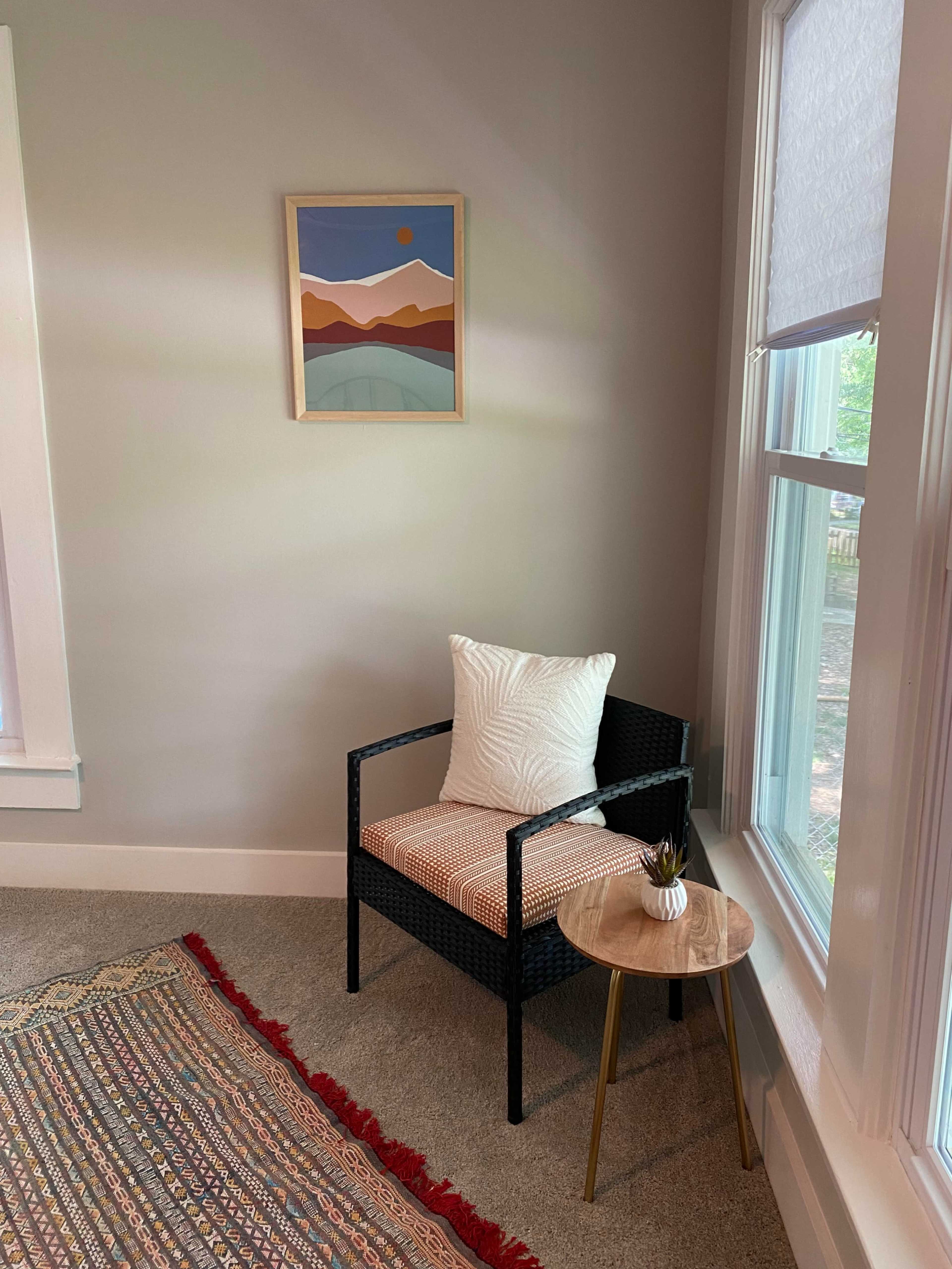 The image shows a small, softly lit corner of a room featuring a chair with a patterned cushion, a side table, and a colorful rug on the floor, with a mountain-themed artwork hanging on the wall.