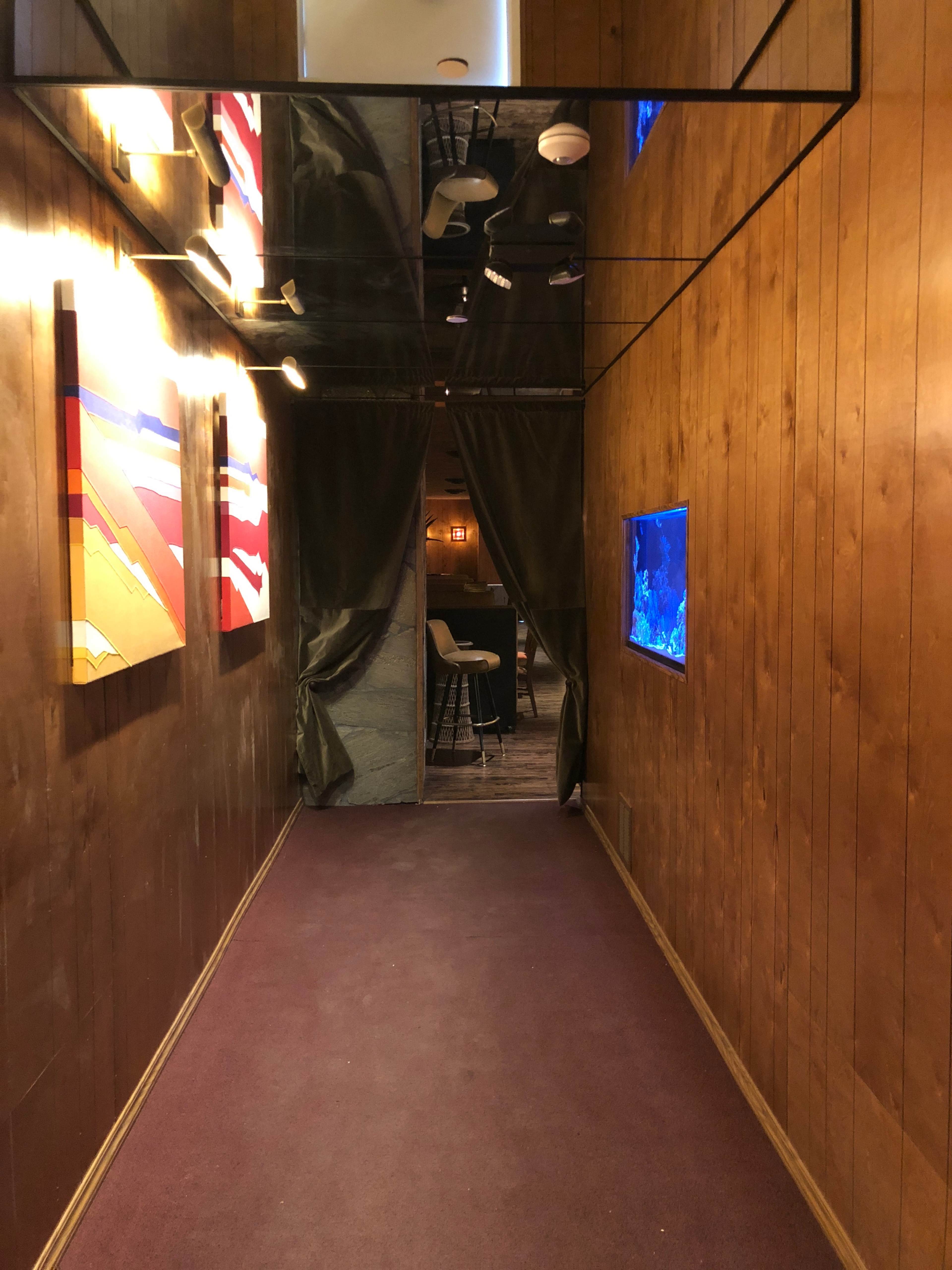 A wooden-paneled hallway features artwork on the walls and leads to a room with an aquarium and a partially opened curtain.