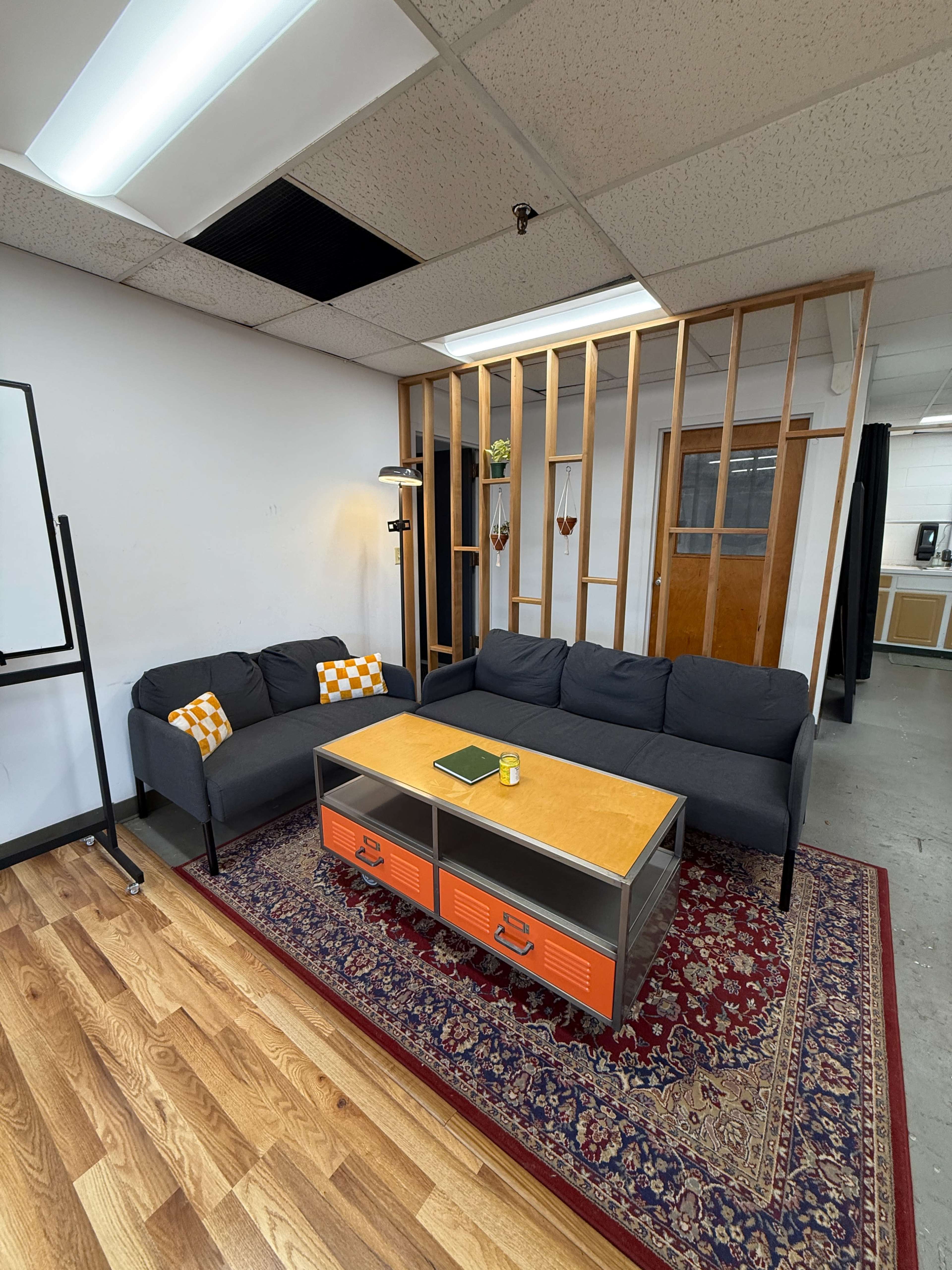 The image shows a modern, furnished workspace featuring a gray sectional sofa, an orange and yellow coffee table, and a decorative wooden partition in a well-lit room.
