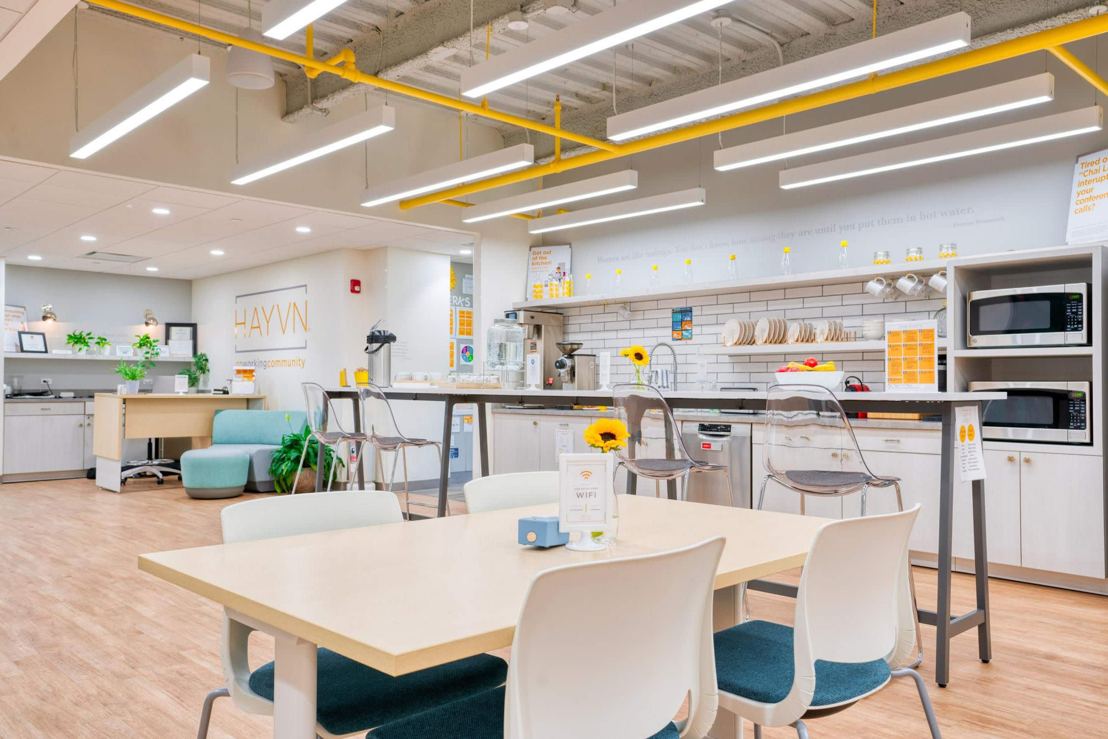 A modern communal kitchen space with bright lighting, a kitchen island, seating area, and various kitchen appliances.