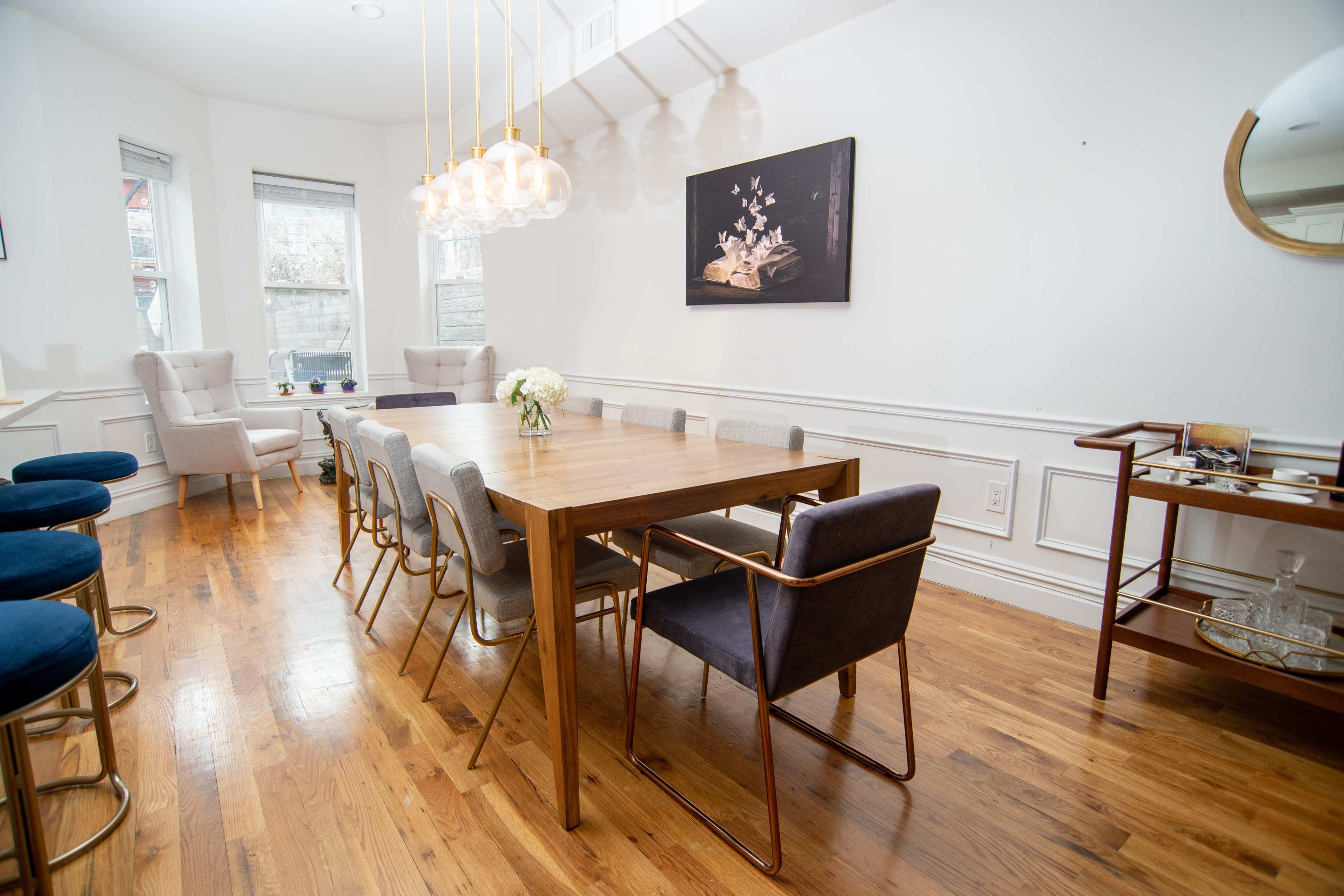 A modern dining room features a wooden table surrounded by upholstered chairs, with a decorative light fixture overhead and a side bar cart against the wall.