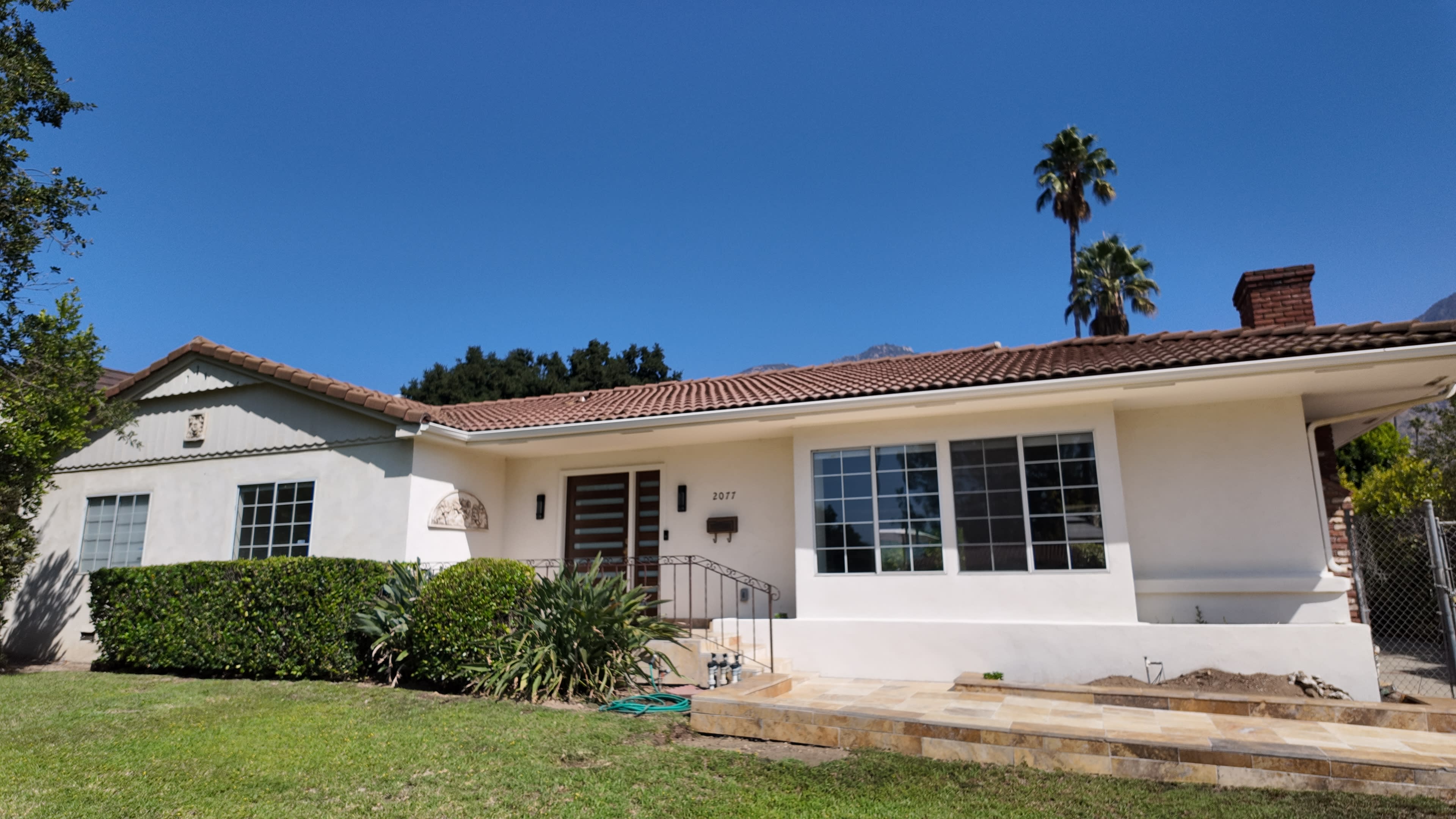 A single-story house with a tiled roof and a green front lawn is shown, featuring large windows and palm trees in the background.