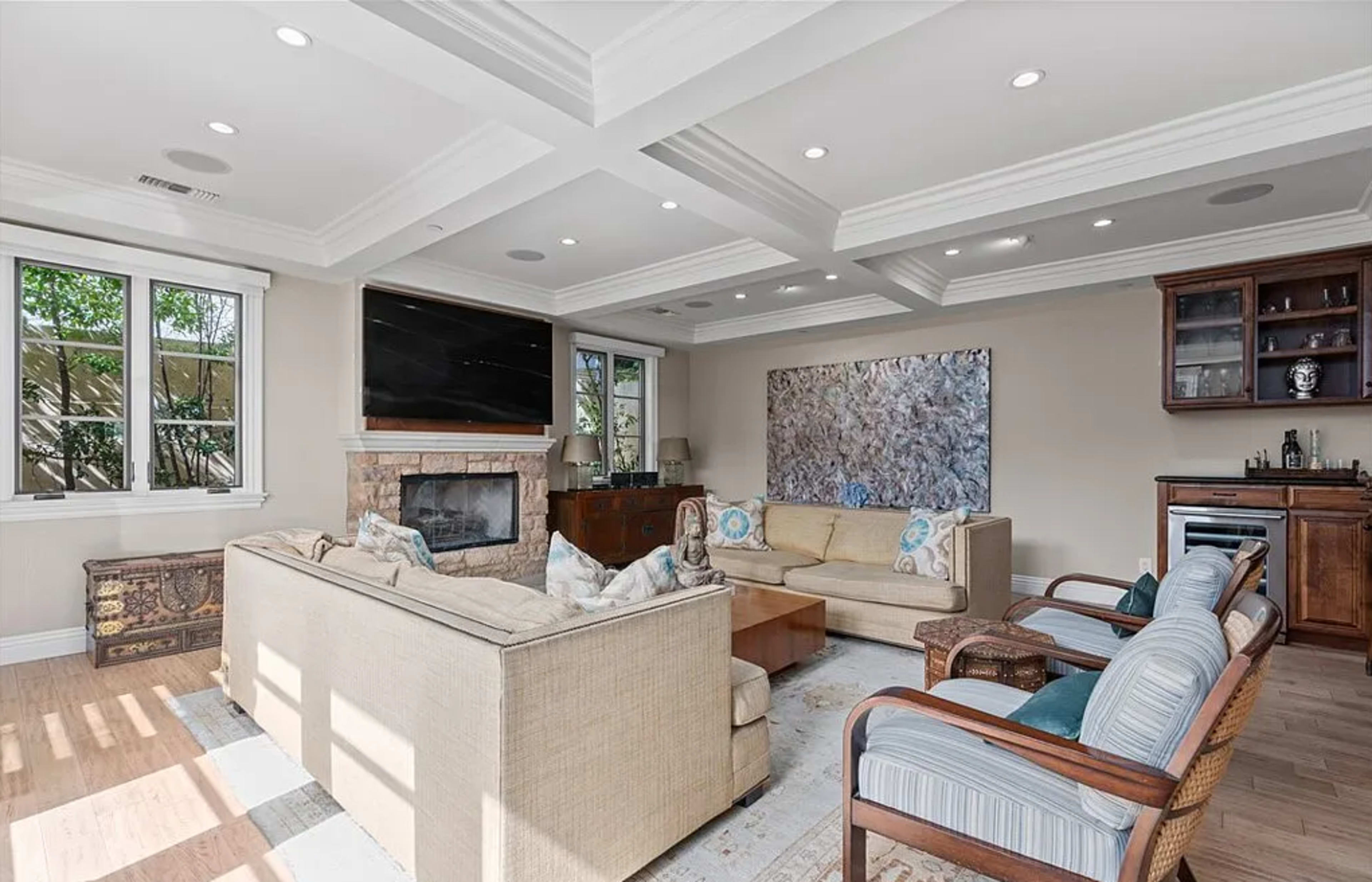 The image shows a modern living room with a beige sectional sofa, wooden accents, and a large wall-mounted TV above a fireplace, complemented by a bar area in the corner.