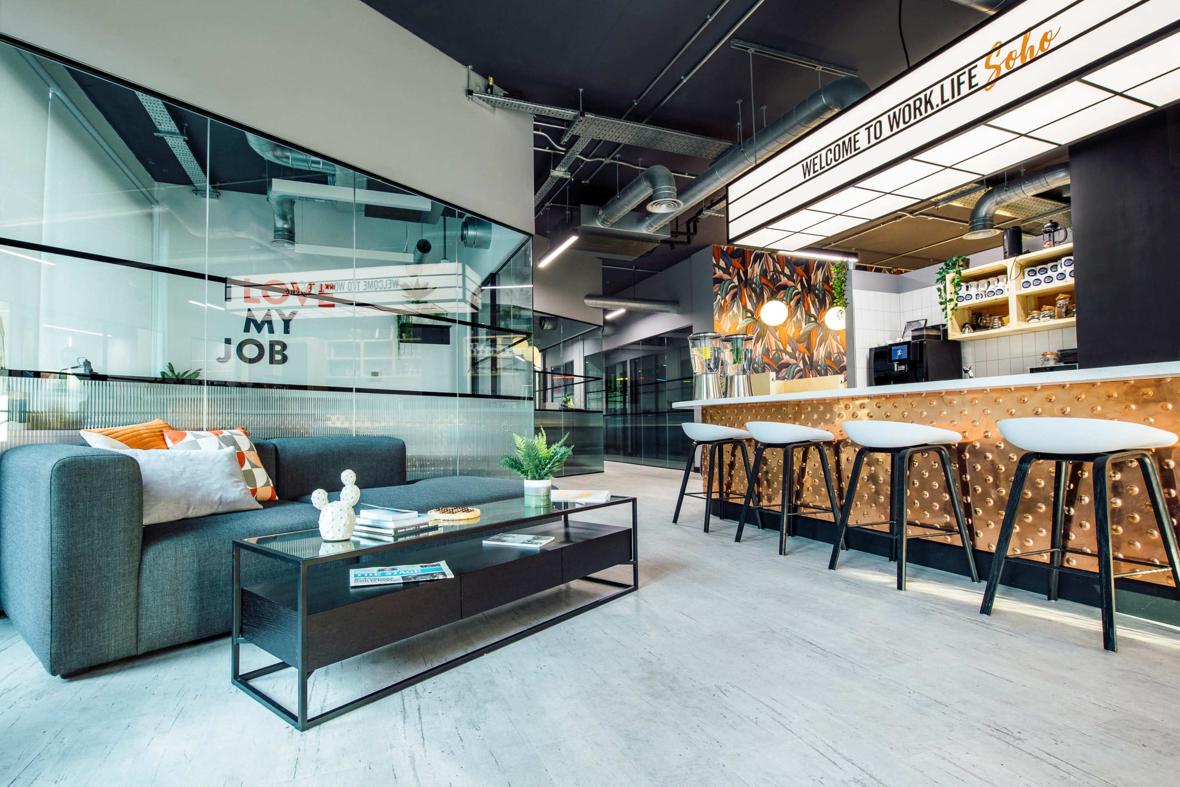 A modern office space featuring a sitting area with a gray sofa, a coffee table, and a bar with stools surrounded by decorative wall art.