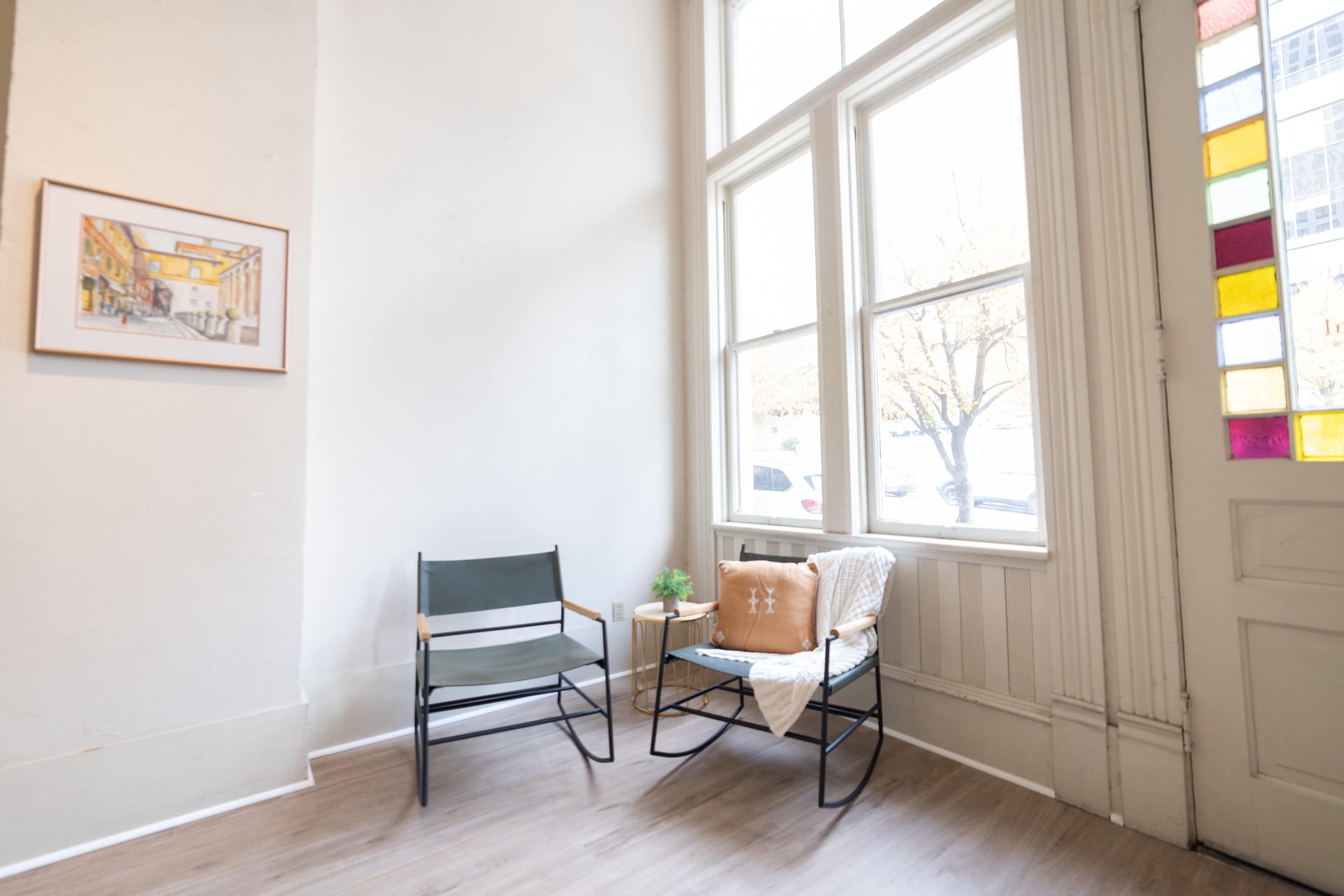 A small seating area features two chairs, a decorative throw, and a plant by a large window with colorful stained glass.