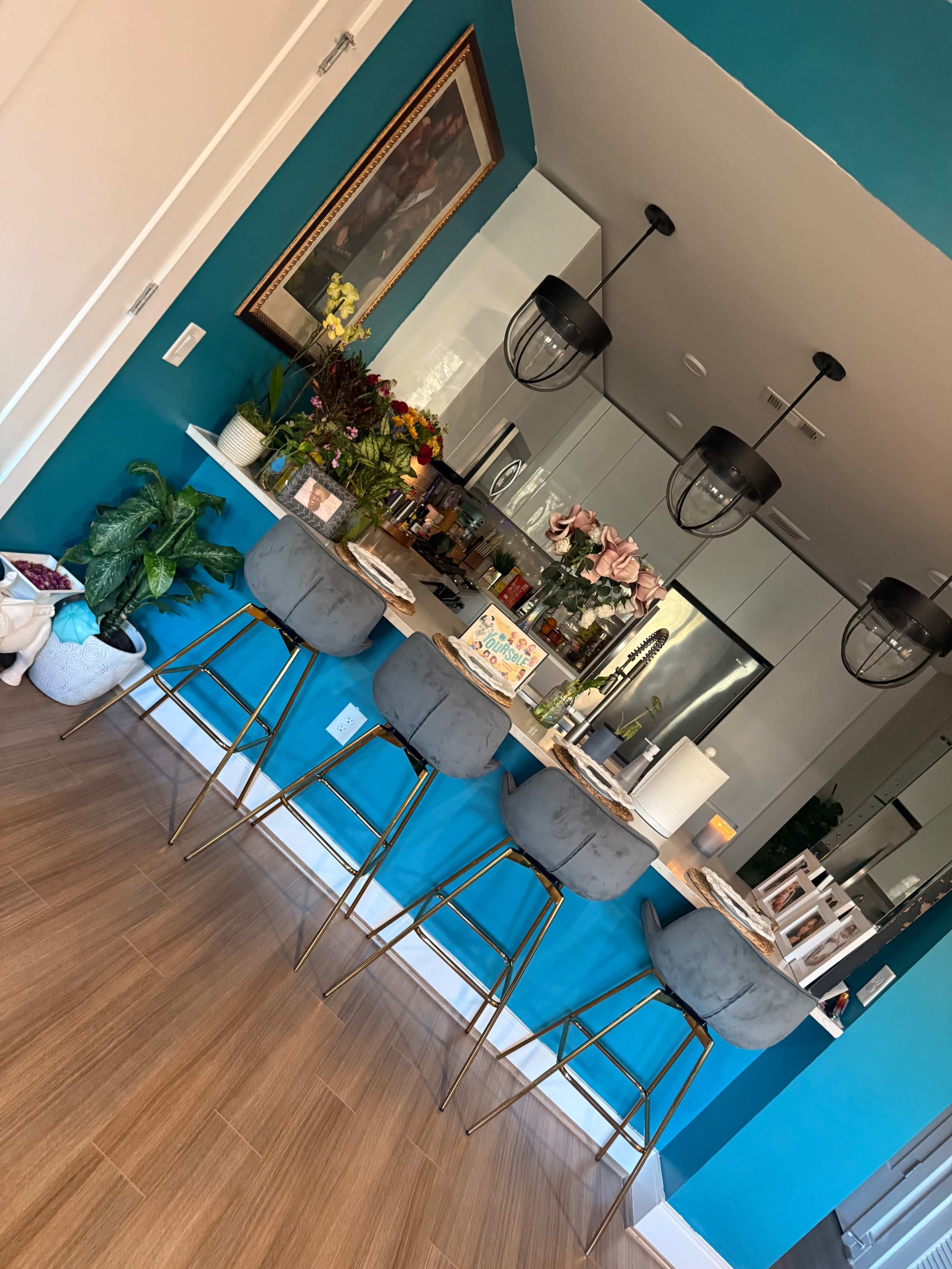A modern kitchen features a turquoise accent wall, a sleek counter with gray bar stools, and various decorative plants and flowers.