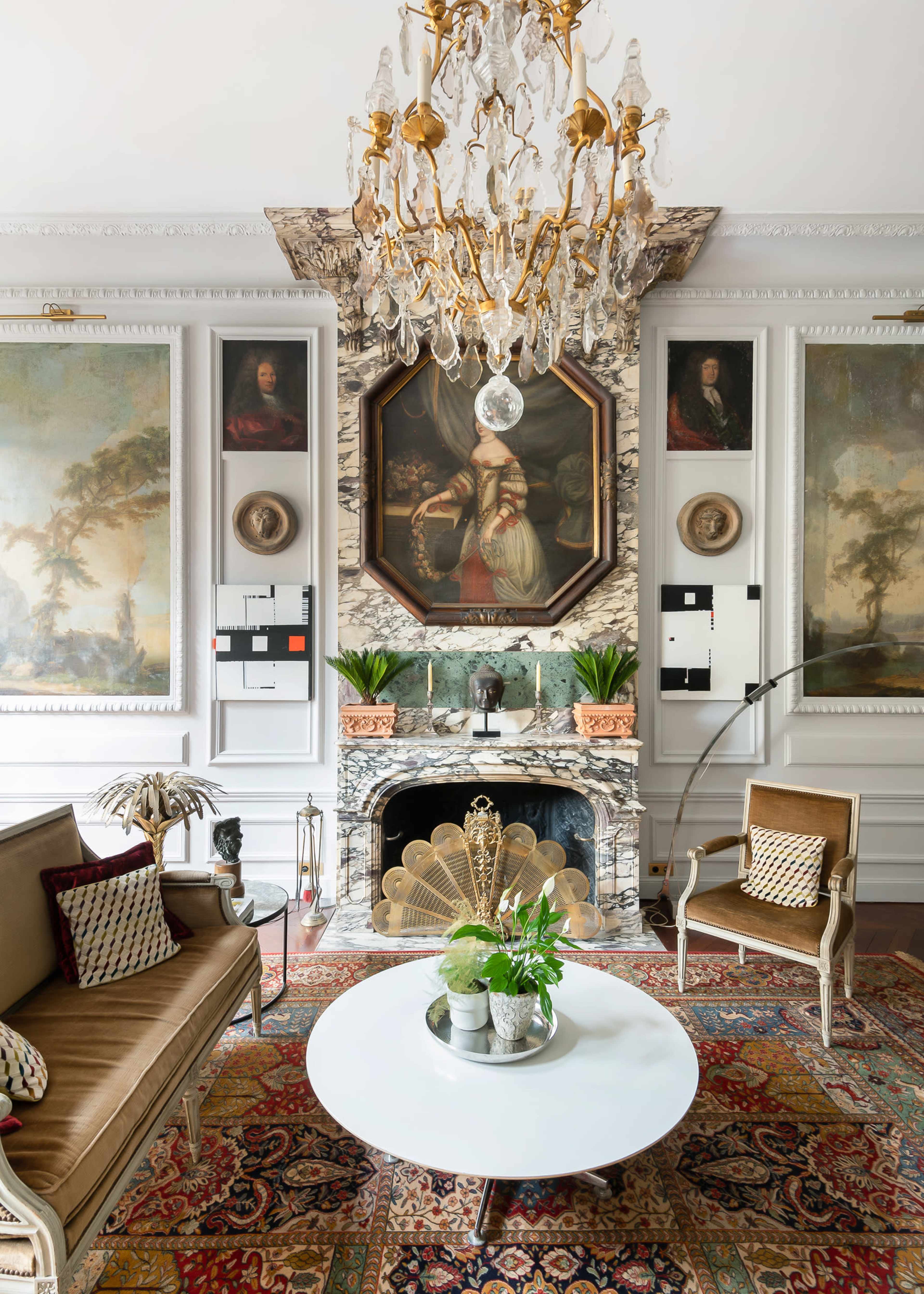 The living room features an ornate chandelier above a round white coffee table, surrounded by upholstered seating and framed artwork, with a decorative fireplace as the focal point.