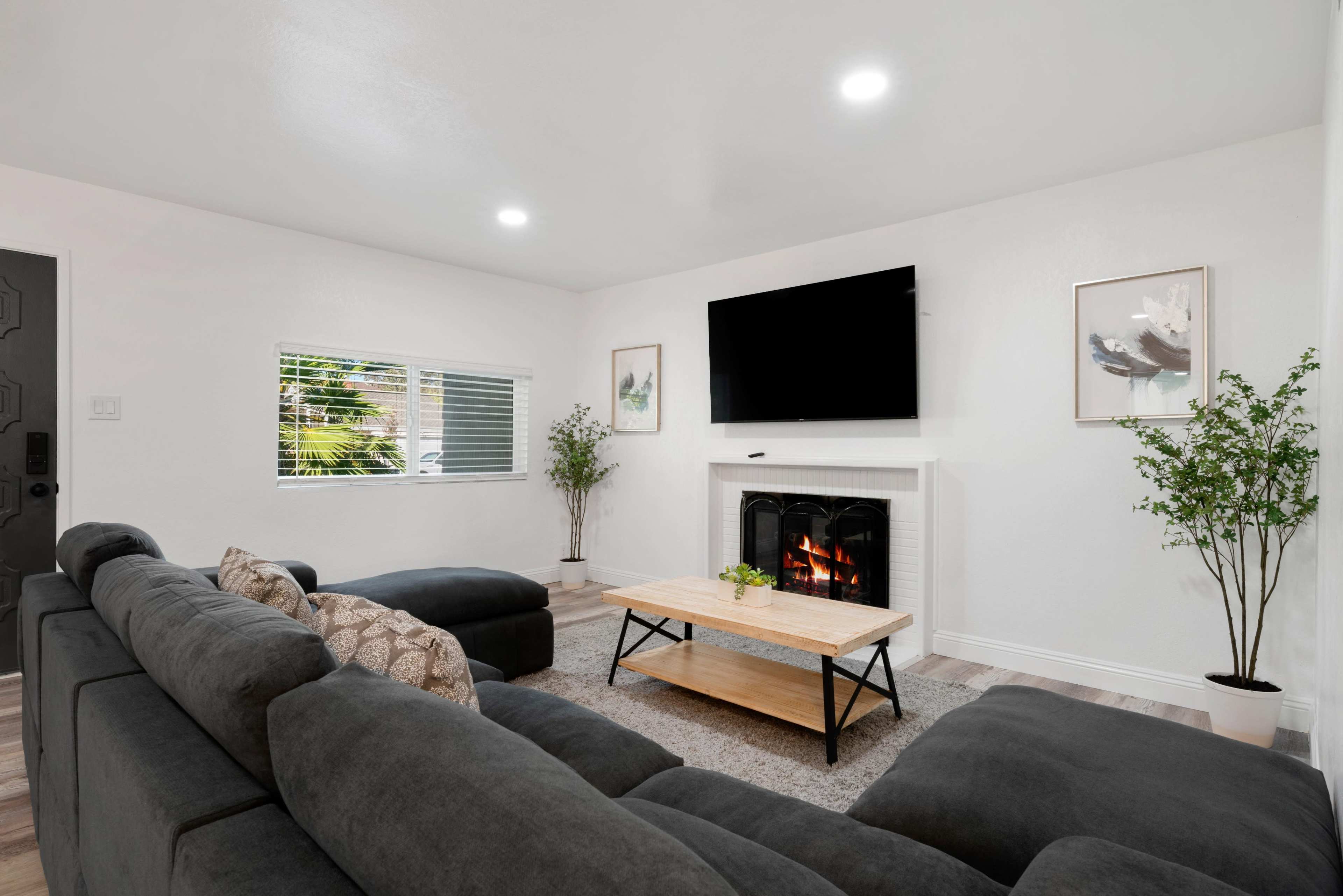 A modern living room features a sectional sofa, a wooden coffee table, a fireplace with a flat-screen TV above it, and plants in the corners.