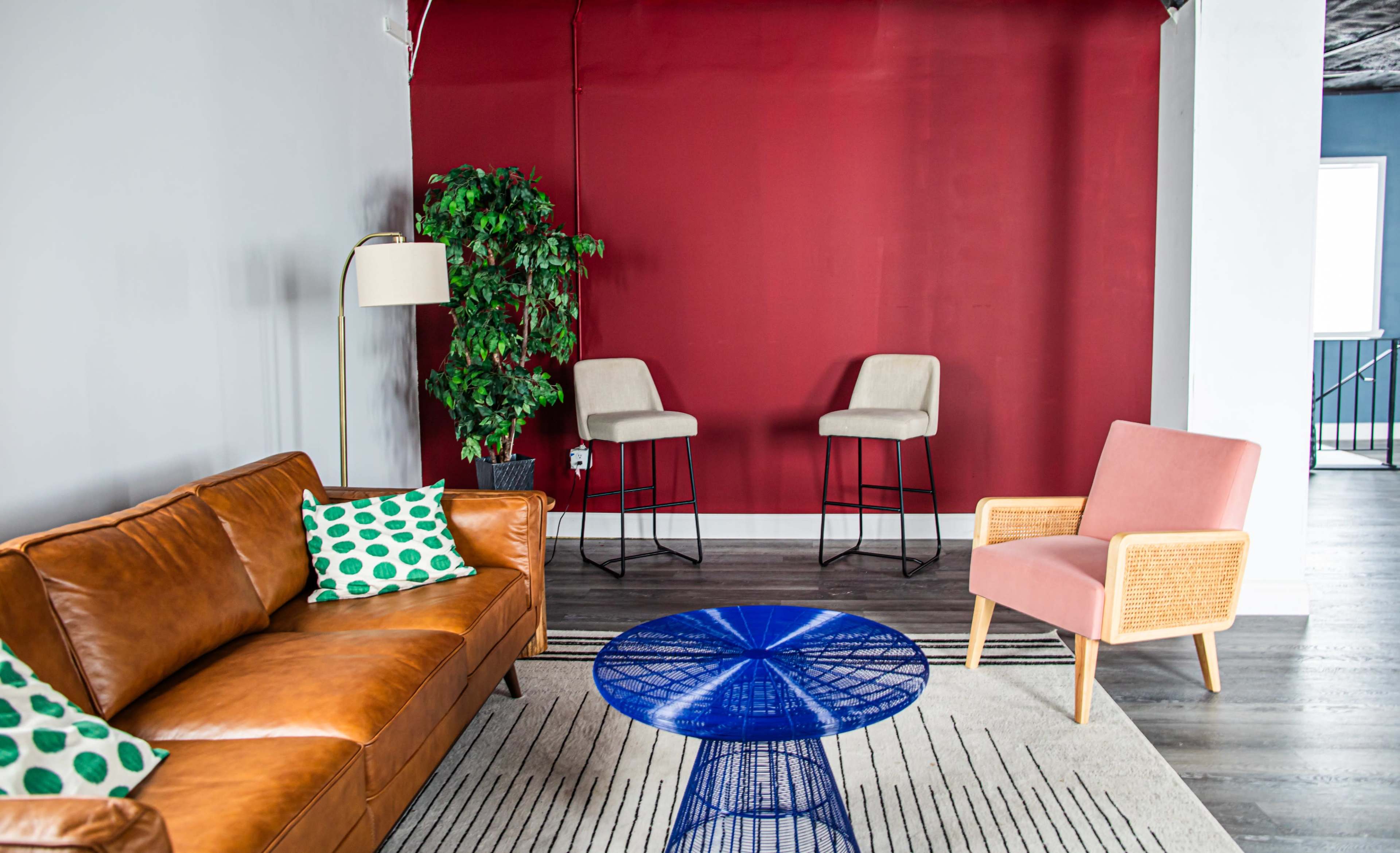 The image shows a seating area with a brown leather sofa, a pink armchair, a blue wire coffee table, and two bar stools against a red wall.