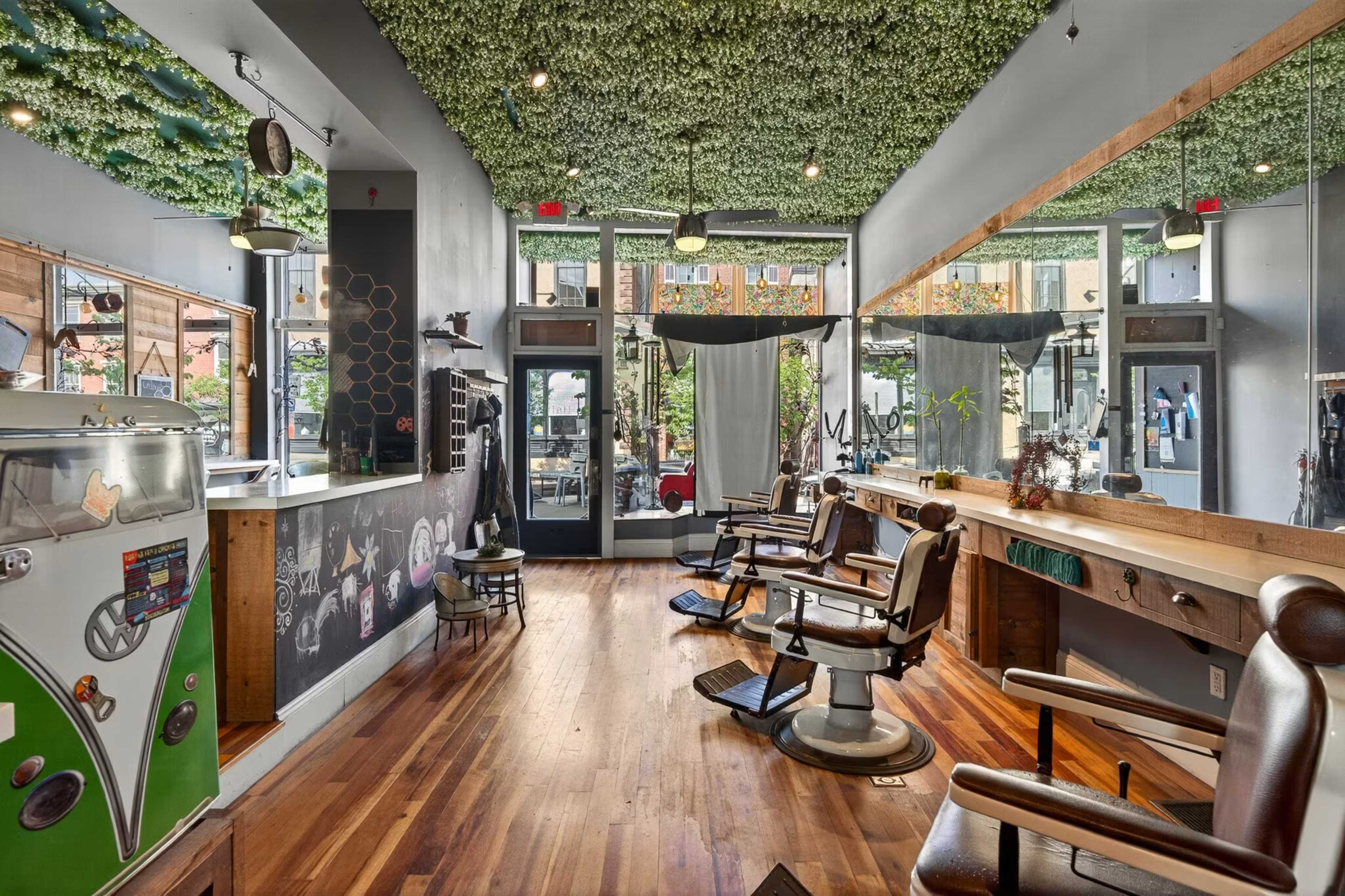 The image shows a modern barbershop with vintage barber chairs, a green foliage ceiling, large mirrors, and an entrance that opens to a city street.