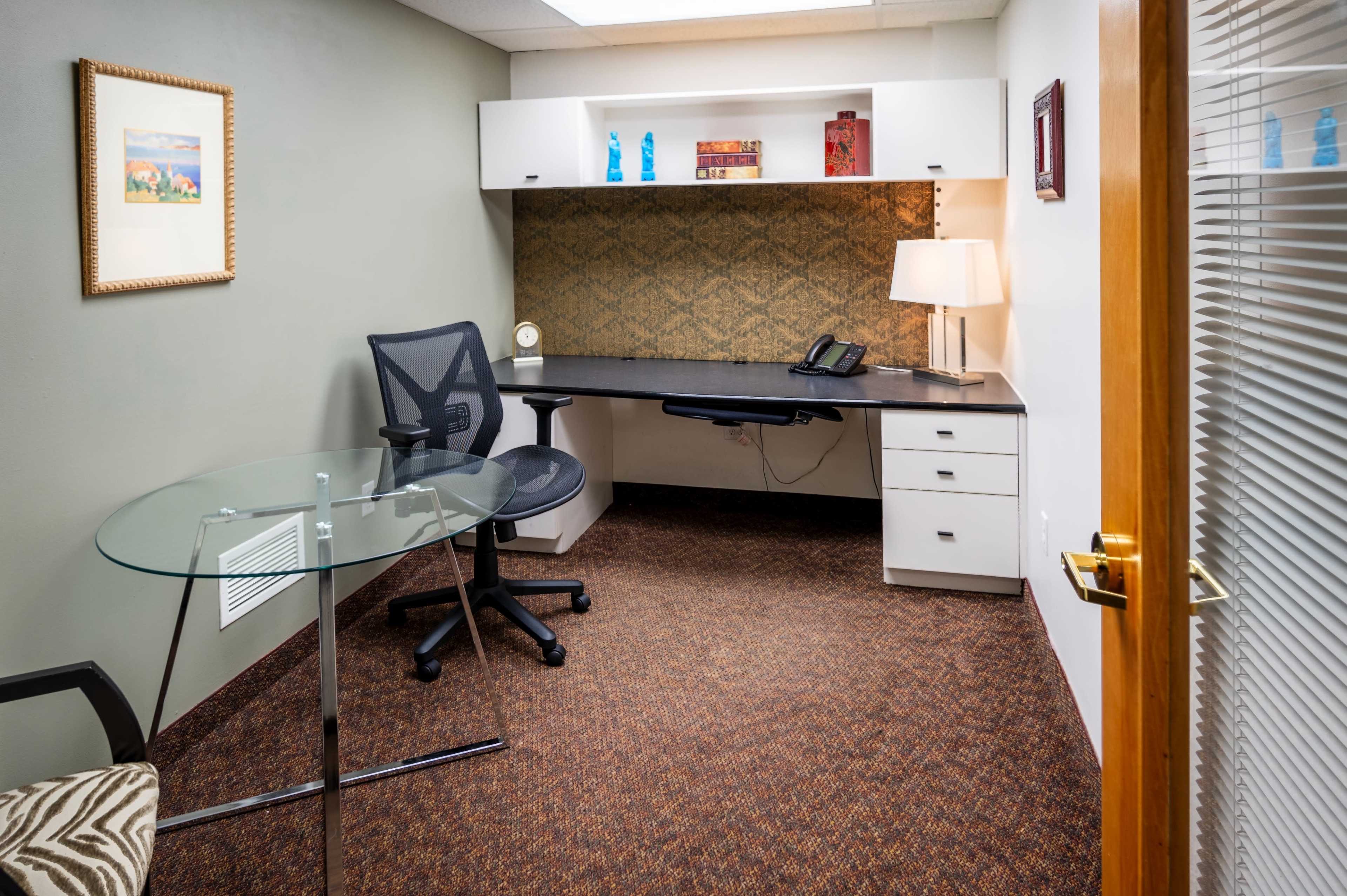 The image shows a small office space featuring a desk with a chair, a glass table, and a decorative wall with a lamp and some items on the shelf.