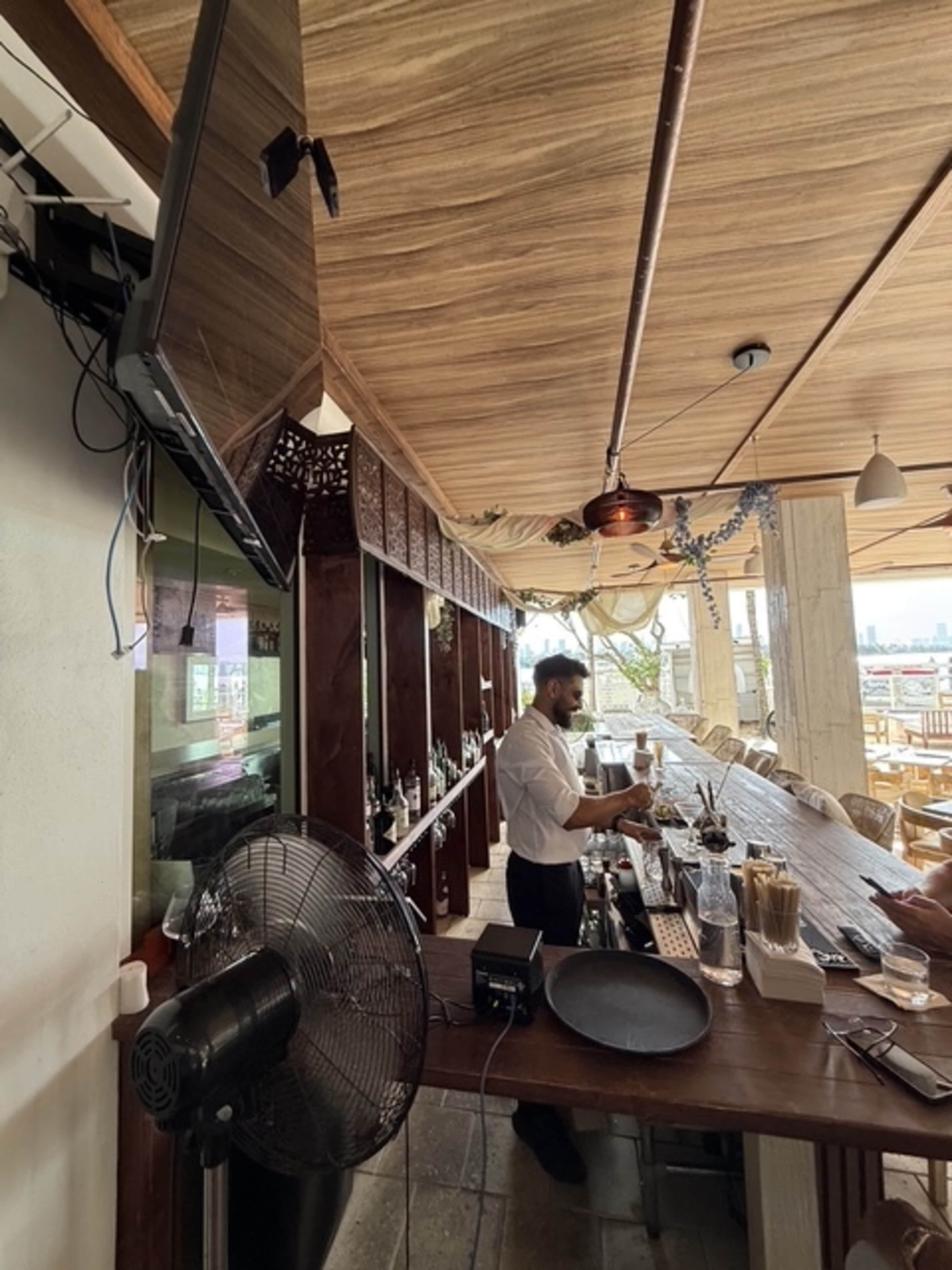 A bartender is preparing drinks at a wooden bar in a restaurant with a fan and a television mounted on the wall.