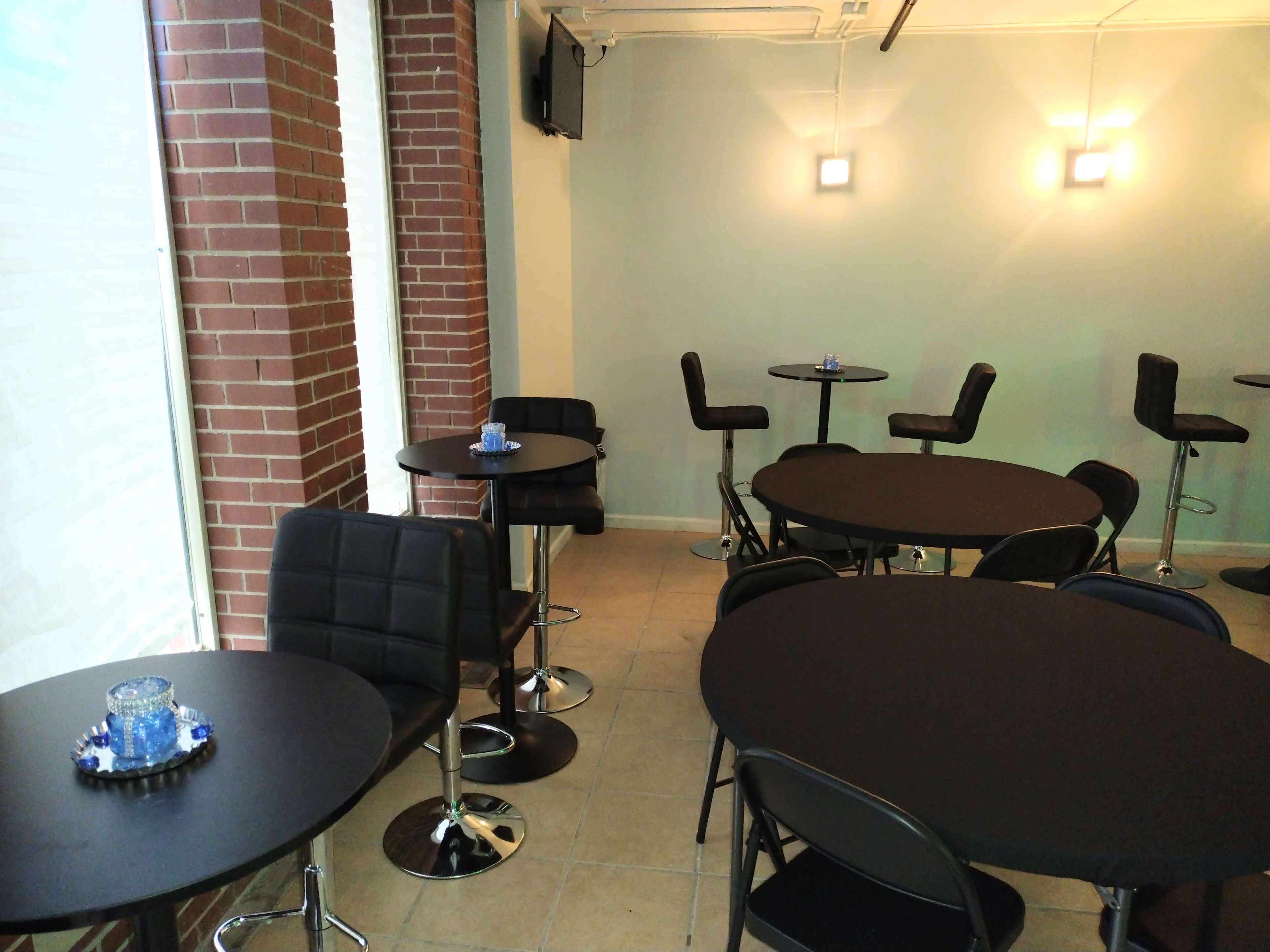 The image depicts a modern café setting with black tables and chairs arranged in a well-lit space featuring brick walls and minimal décor.