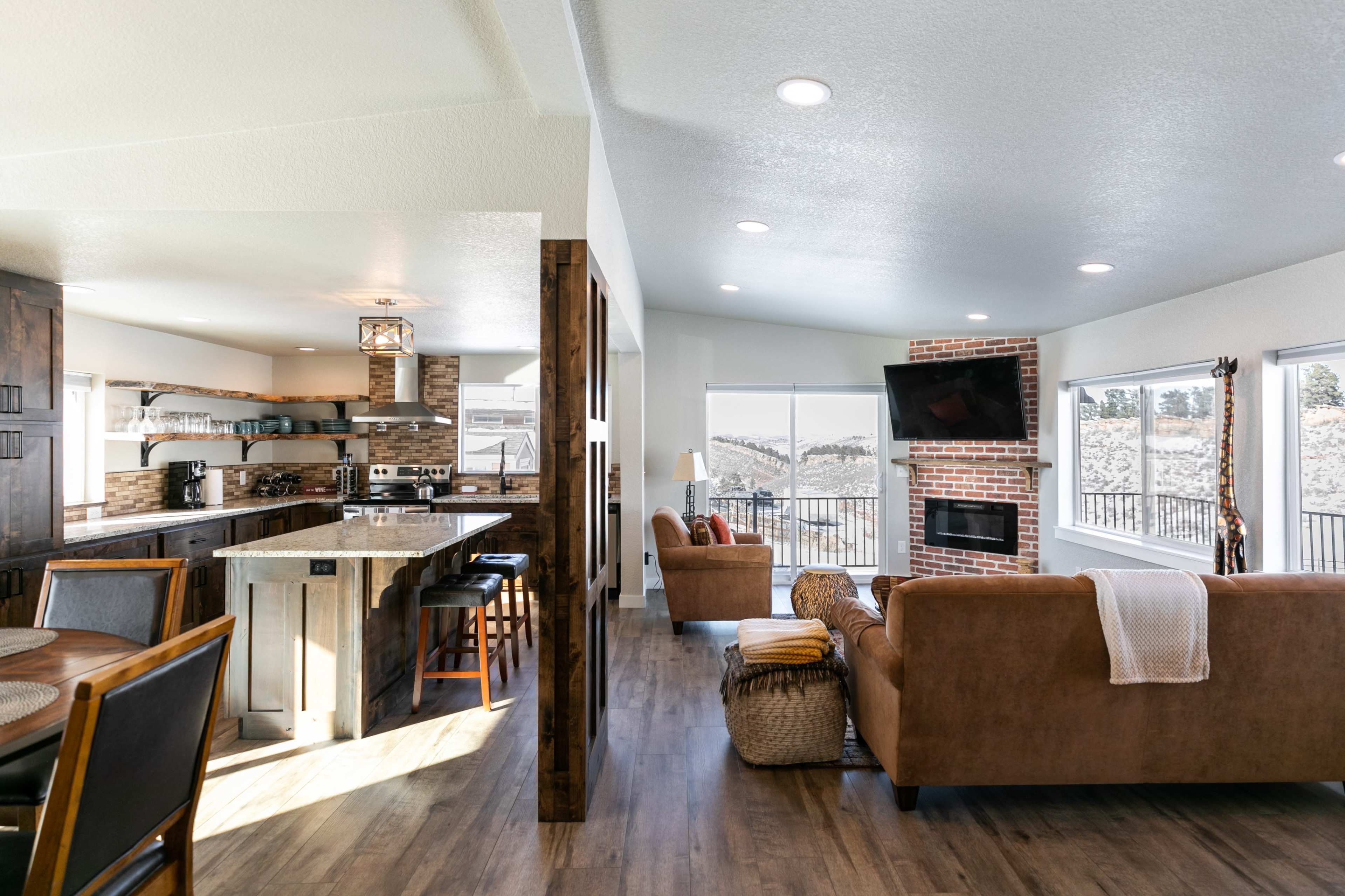 A modern, open-concept living space with a kitchen featuring a marble island, a dining area, and a cozy seating area with a fireplace and large windows overlooking a snowy landscape.