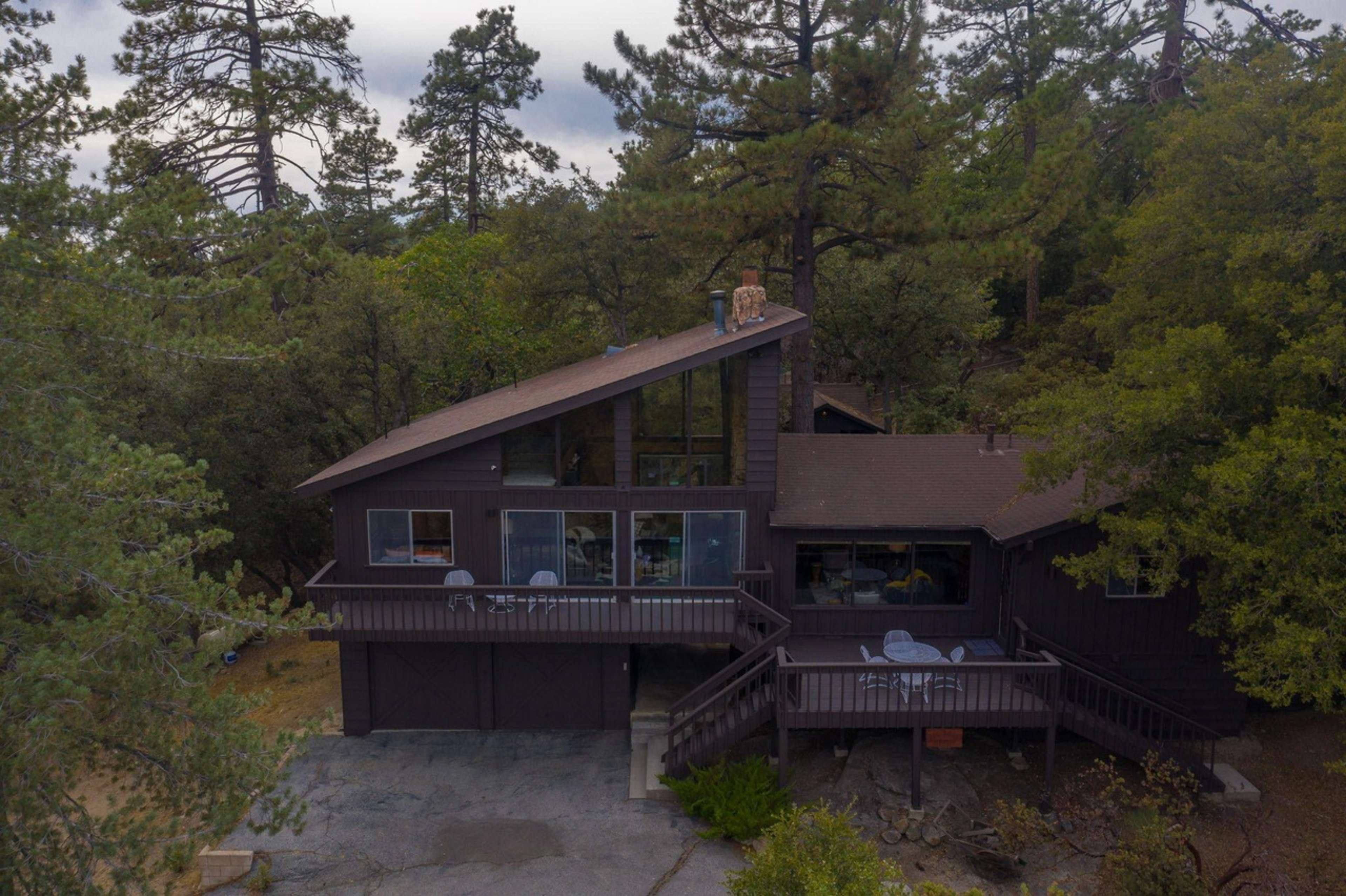 A two-story dark wooden house is nestled among tall trees in a wooded area.