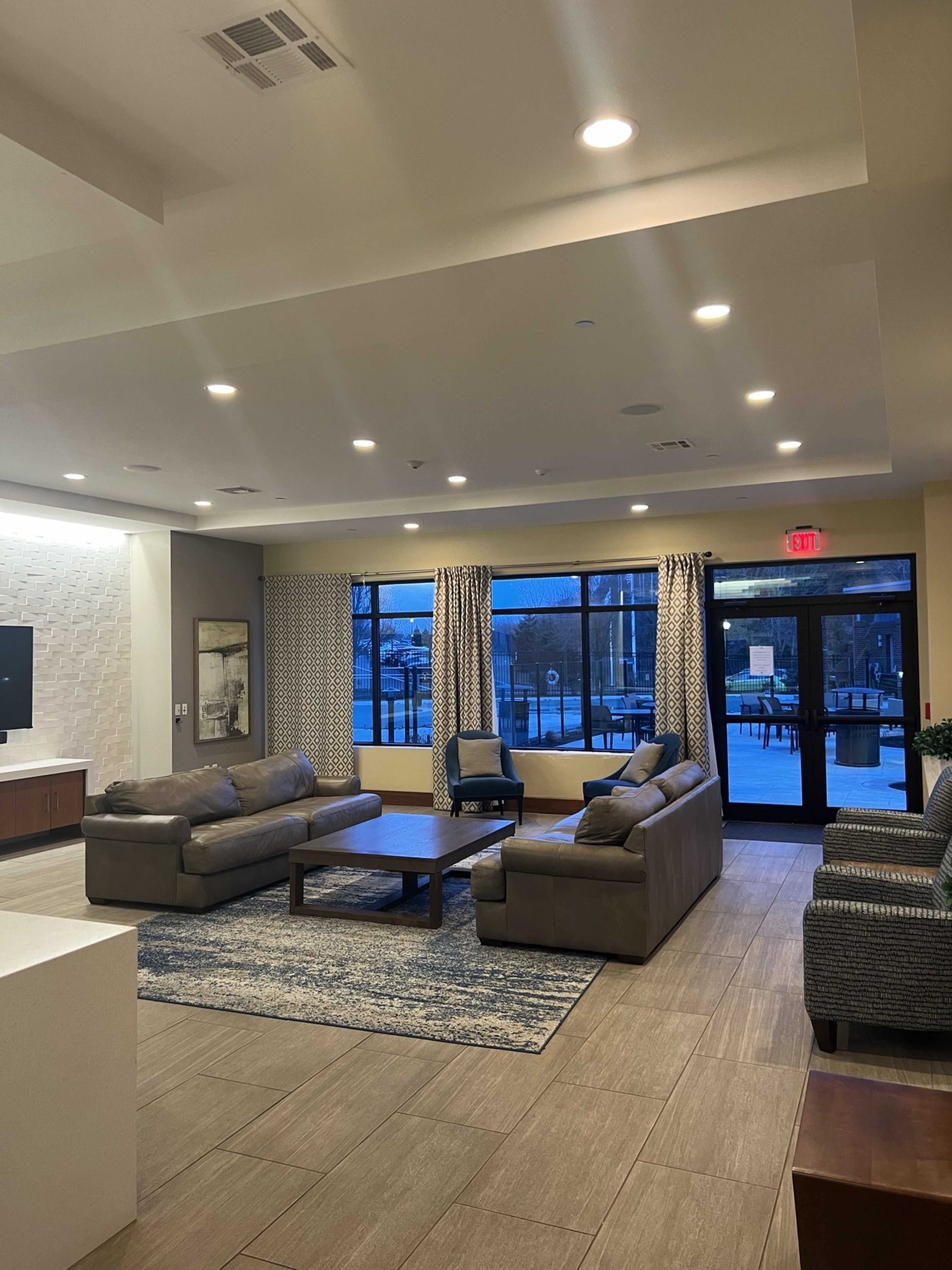 The image shows a modern lobby area featuring two sofas, a coffee table, and large windows with curtains, illuminating the space with natural light.