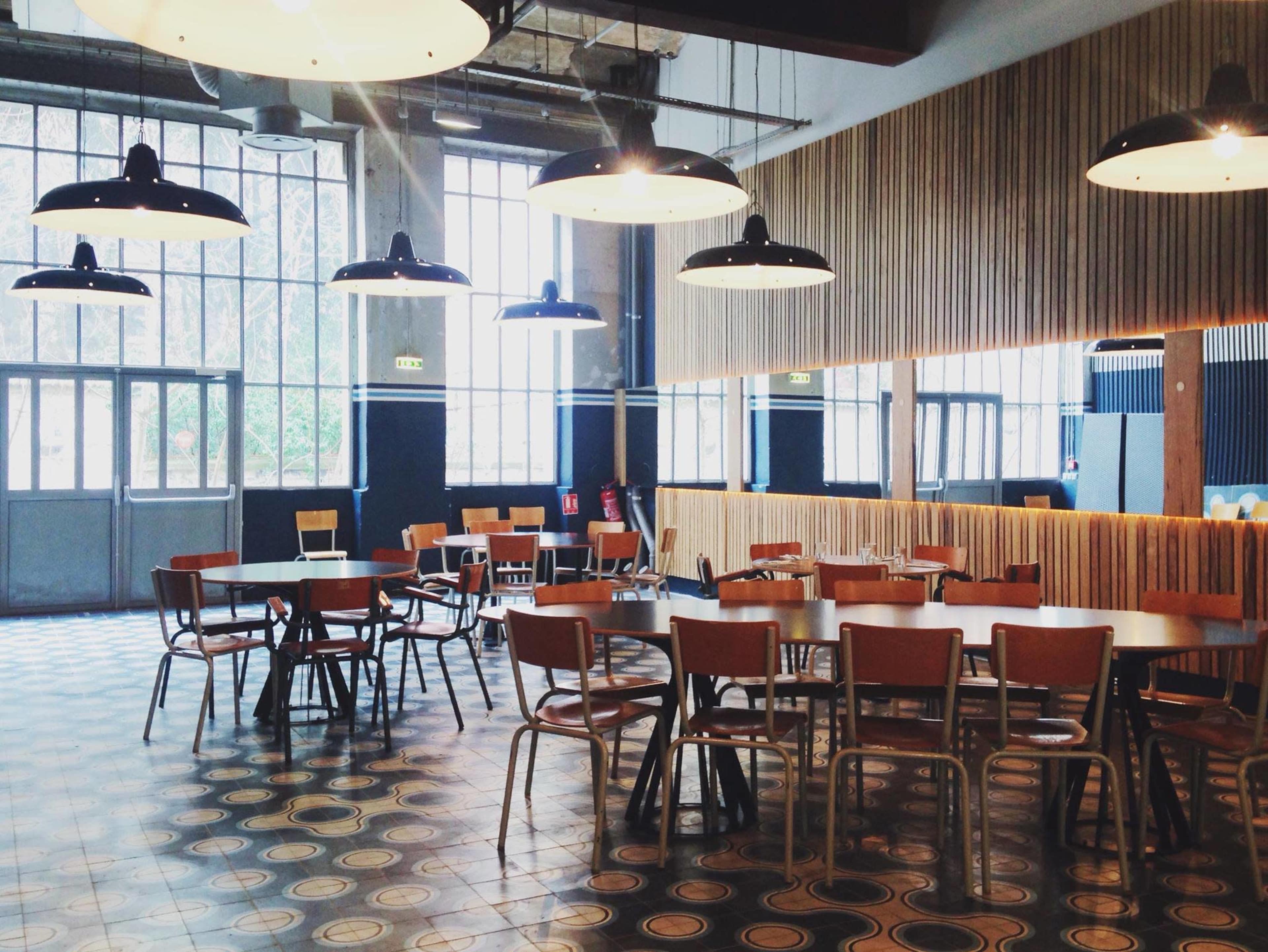 A spacious, modern café interior with several wooden tables and chairs, large windows allowing natural light, and industrial-style lighting fixtures.
