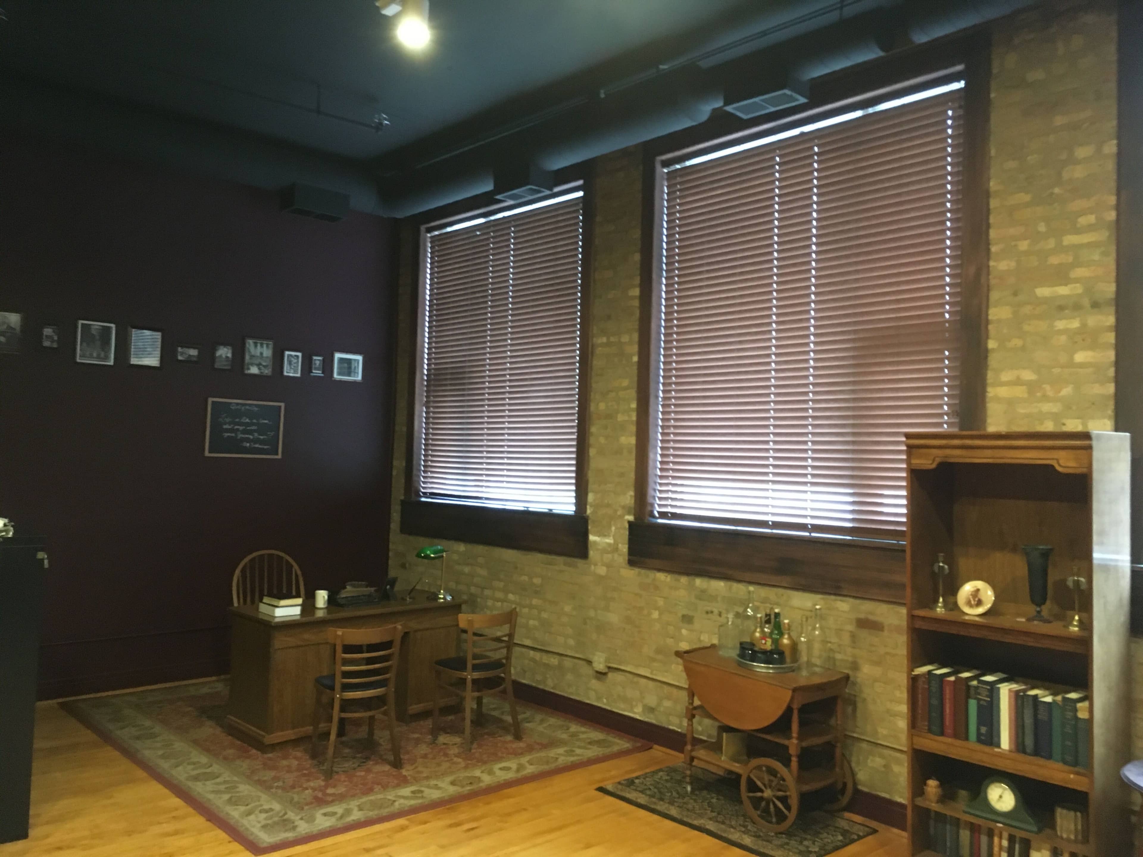 The image shows a sparsely furnished office space with a wooden desk, a chair, and two large windows covered with blinds, against a backdrop of exposed brick walls.
