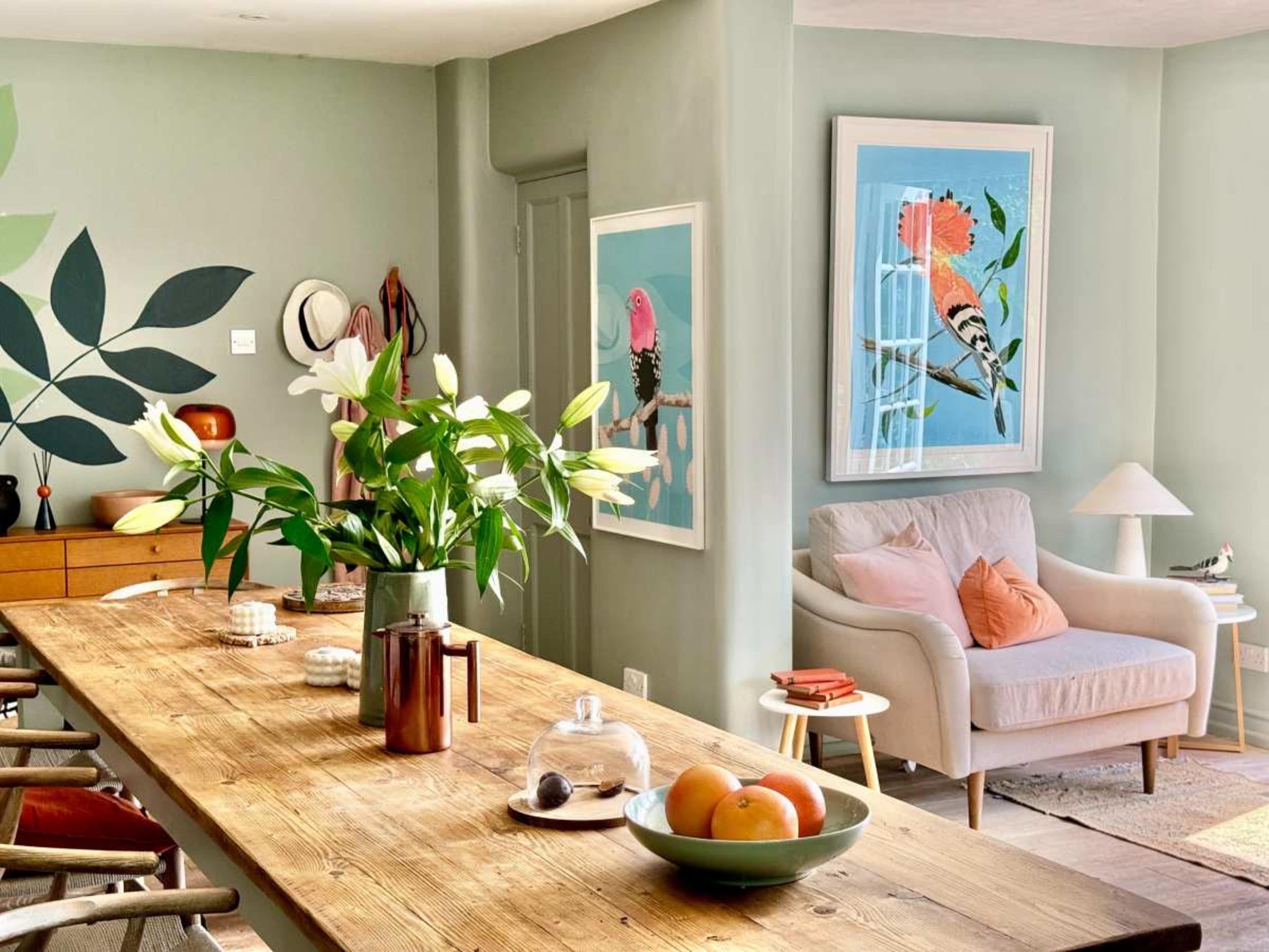 A modern living space features a large wooden dining table, framed artwork on the walls, and a cozy armchair with decorative pillows.