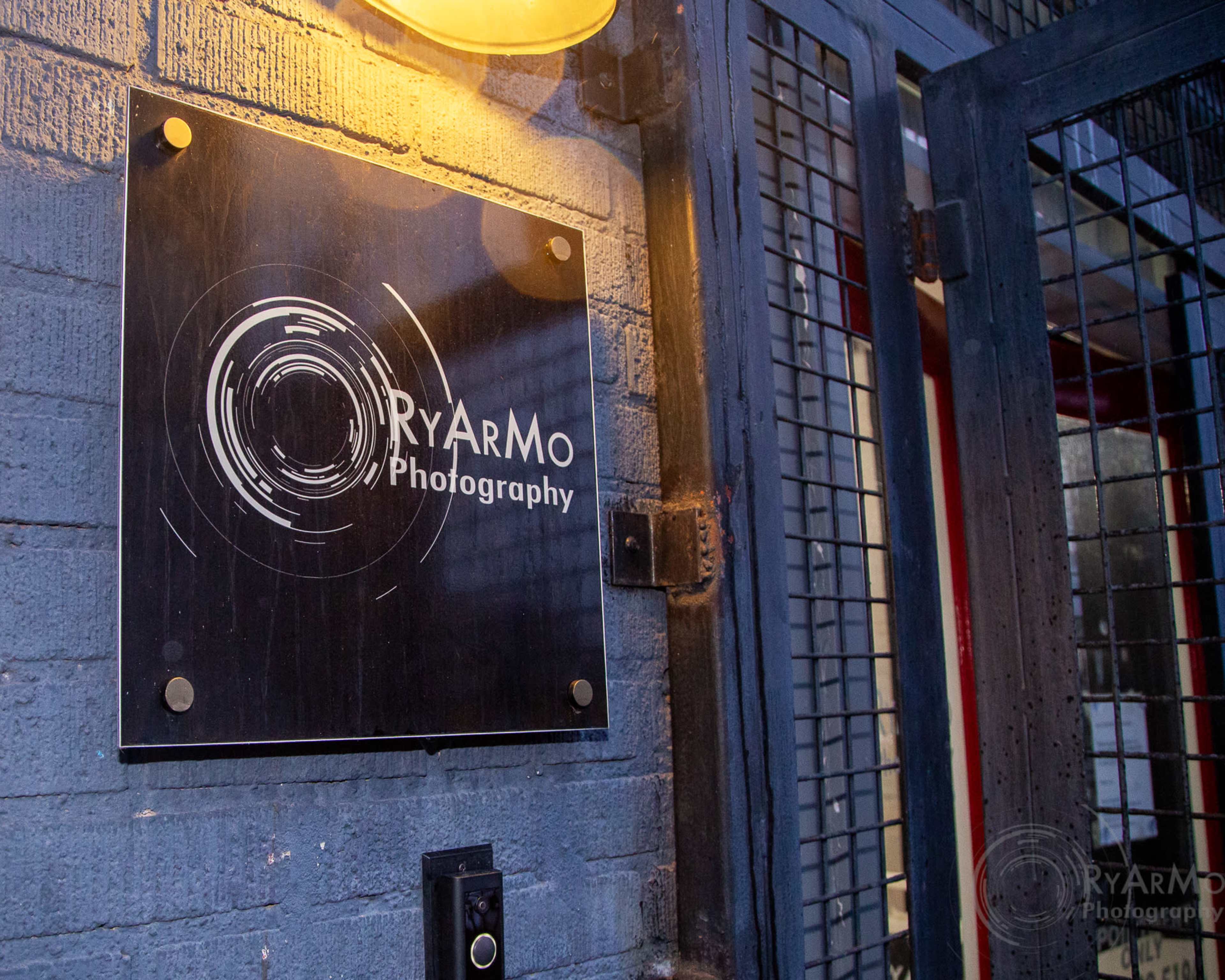 A sign for RYARMO Photography is mounted on a brick wall next to a gated entrance.