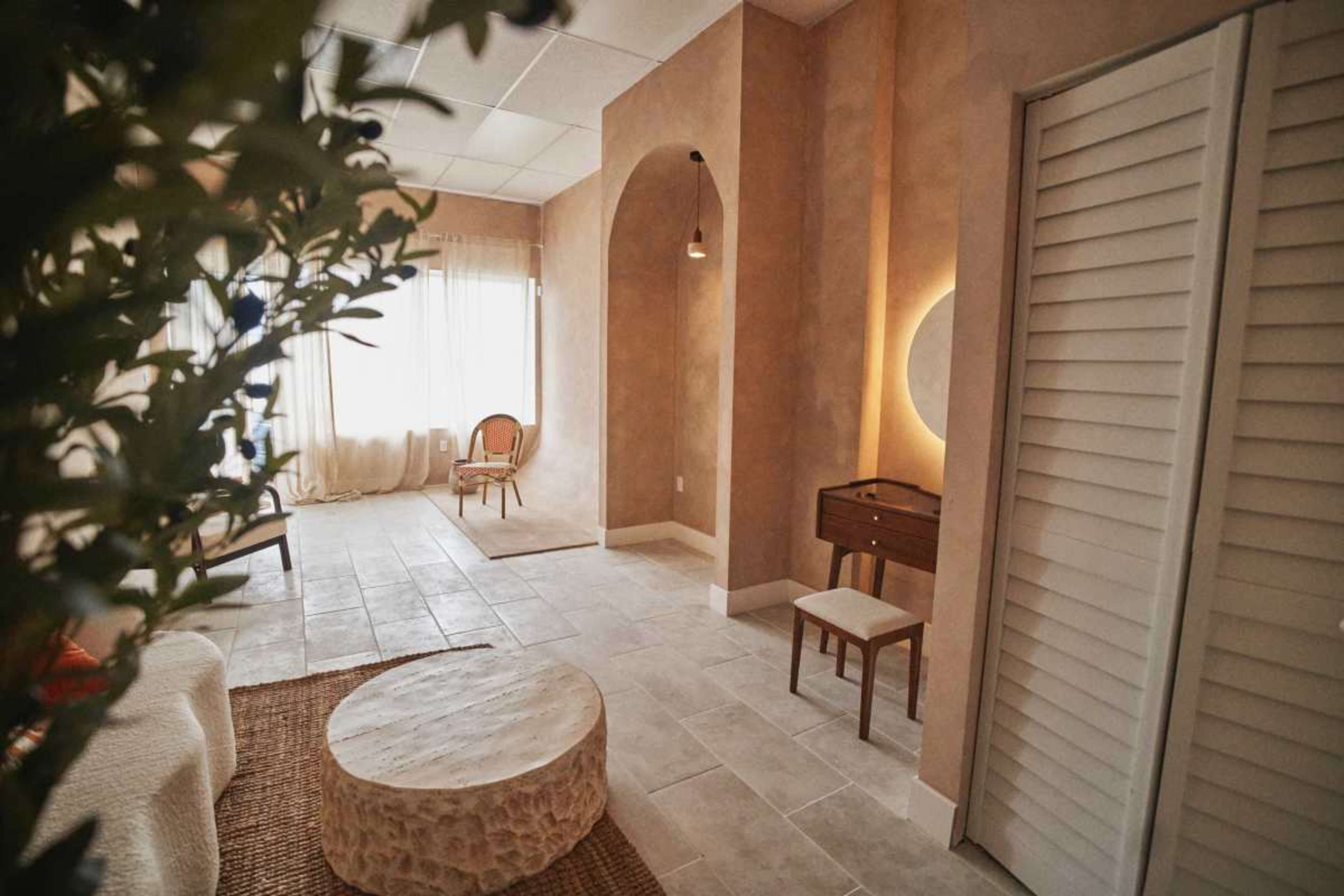 The image shows a softly lit room with neutral-colored walls, a round coffee table, a small wooden desk, and a chair, complemented by natural light filtering through sheer curtains.