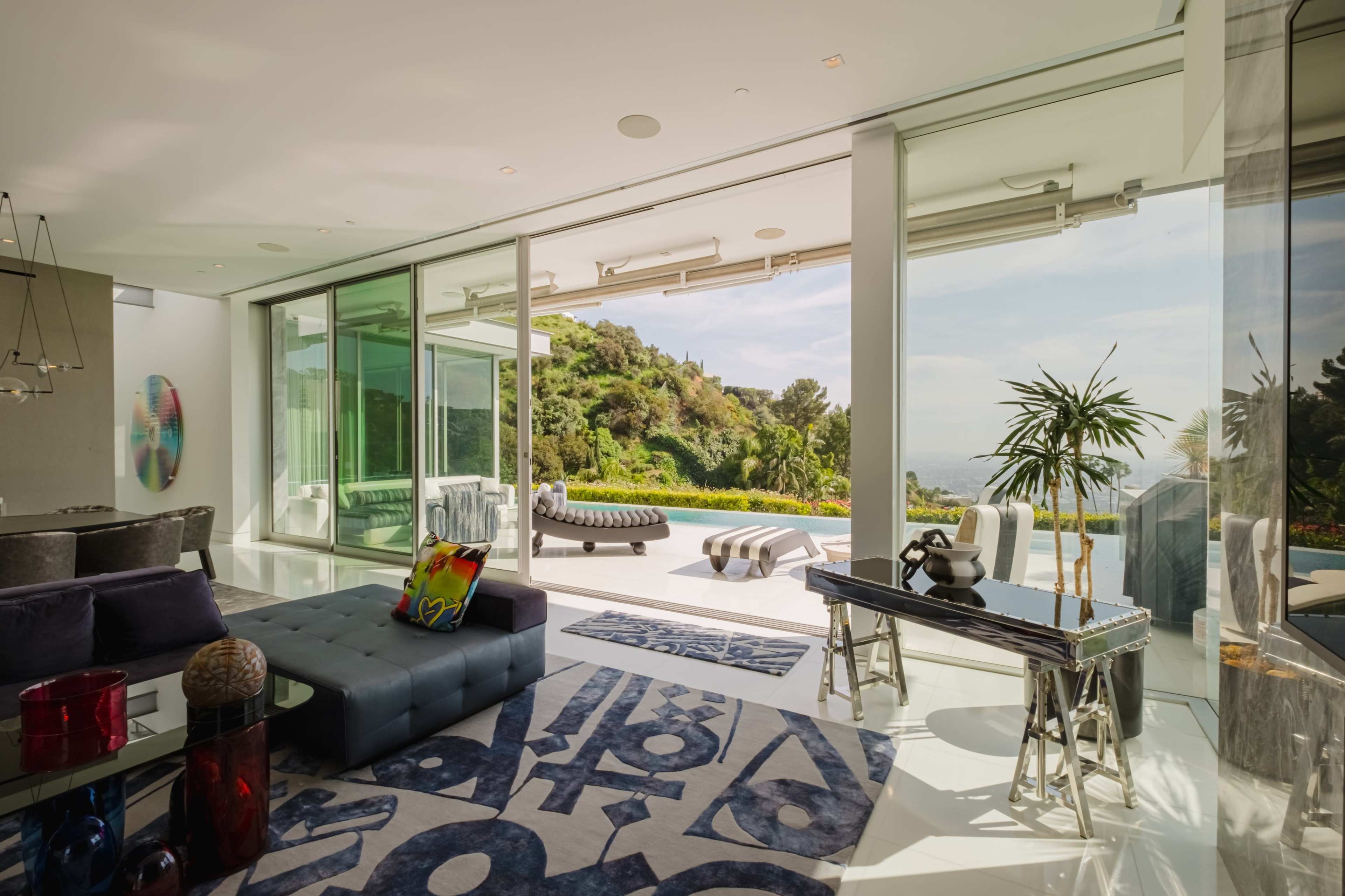 The image shows a modern living room with large glass walls that open to a scenic view of greenery and hills.