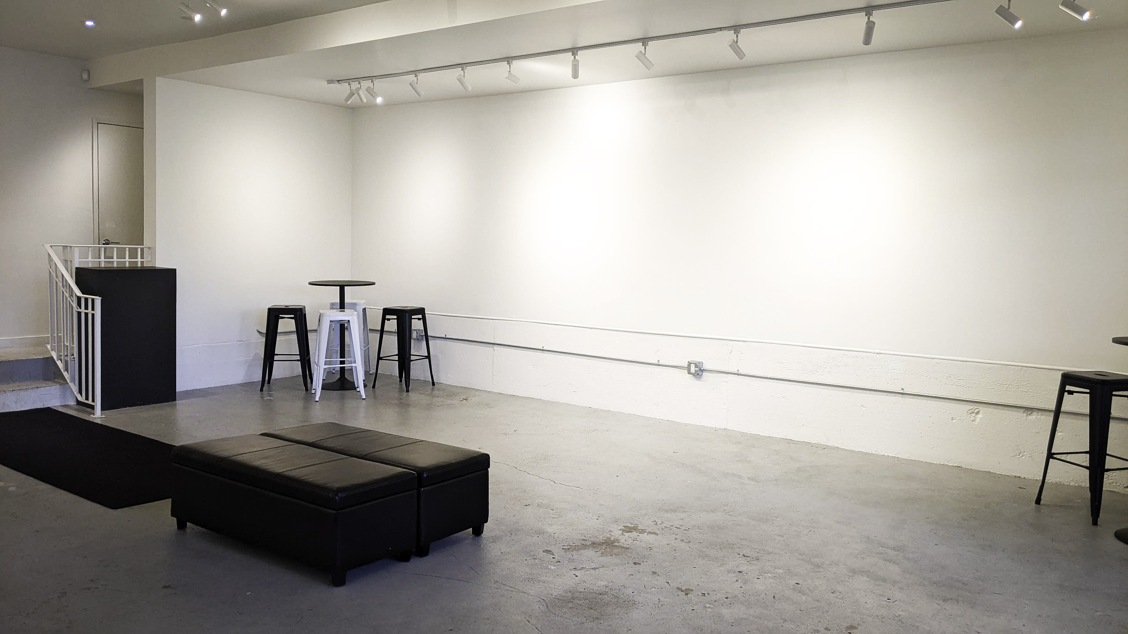 A minimalist room with a concrete floor, two high tables, and four stools against a blank white wall.