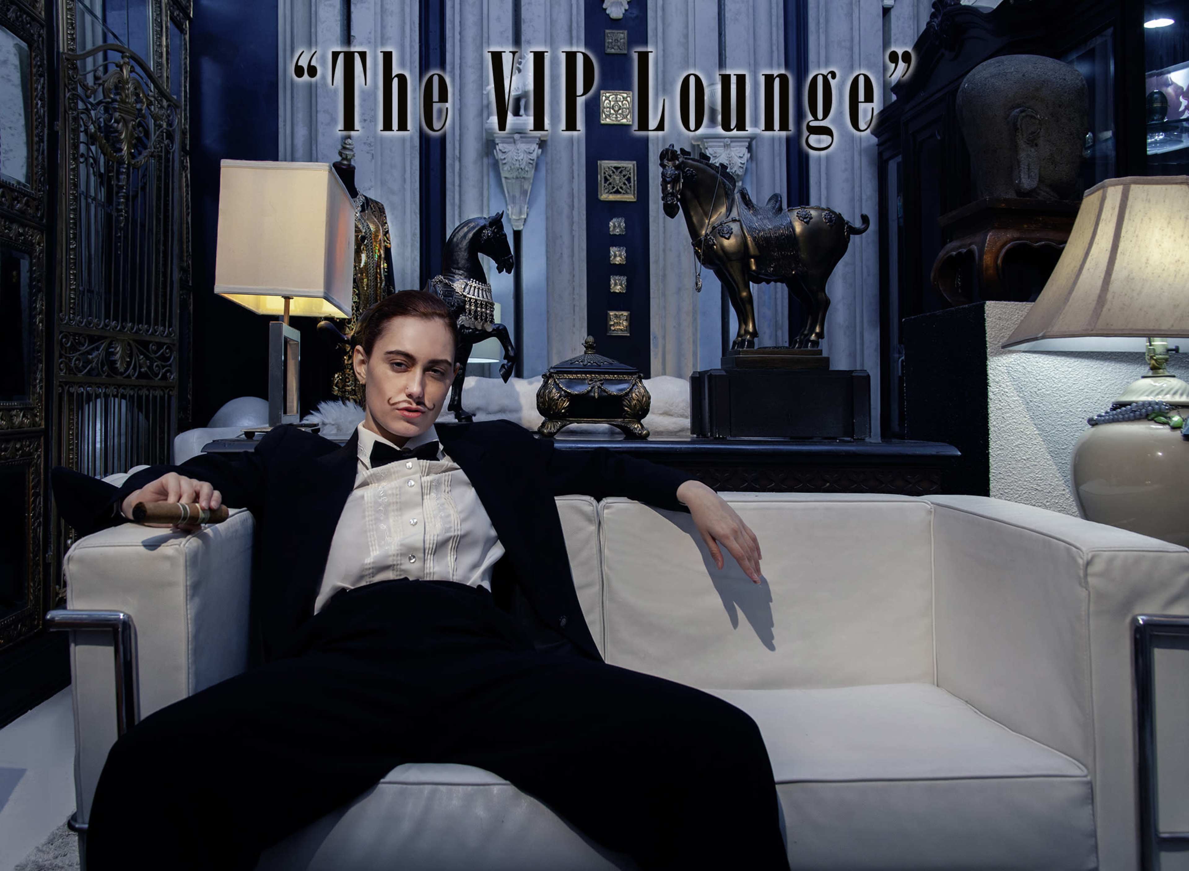 A person in formal attire sits relaxed on a white couch in a stylishly decorated lounge surrounded by various decorative items.