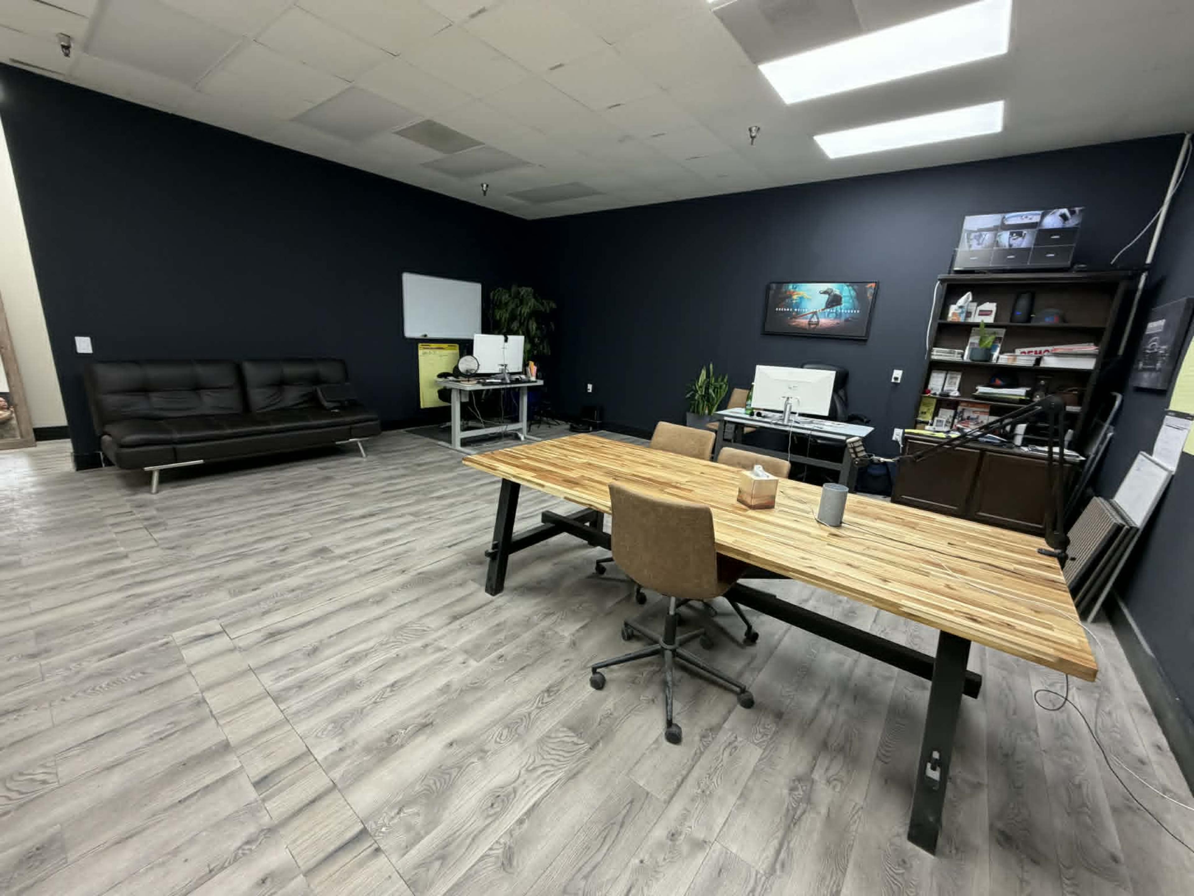 The image shows a spacious office with a large wooden table, a chair, a sofa, and shelving against a dark wall.